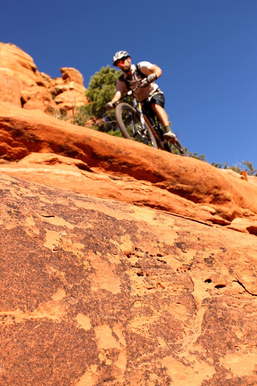 A mountain biker navigating a rocky terrain with vibrant red rock formations in the background under a clear blue sky. The focus is on the textured foreground rock as the biker approaches. Hangover mountain bike trail.