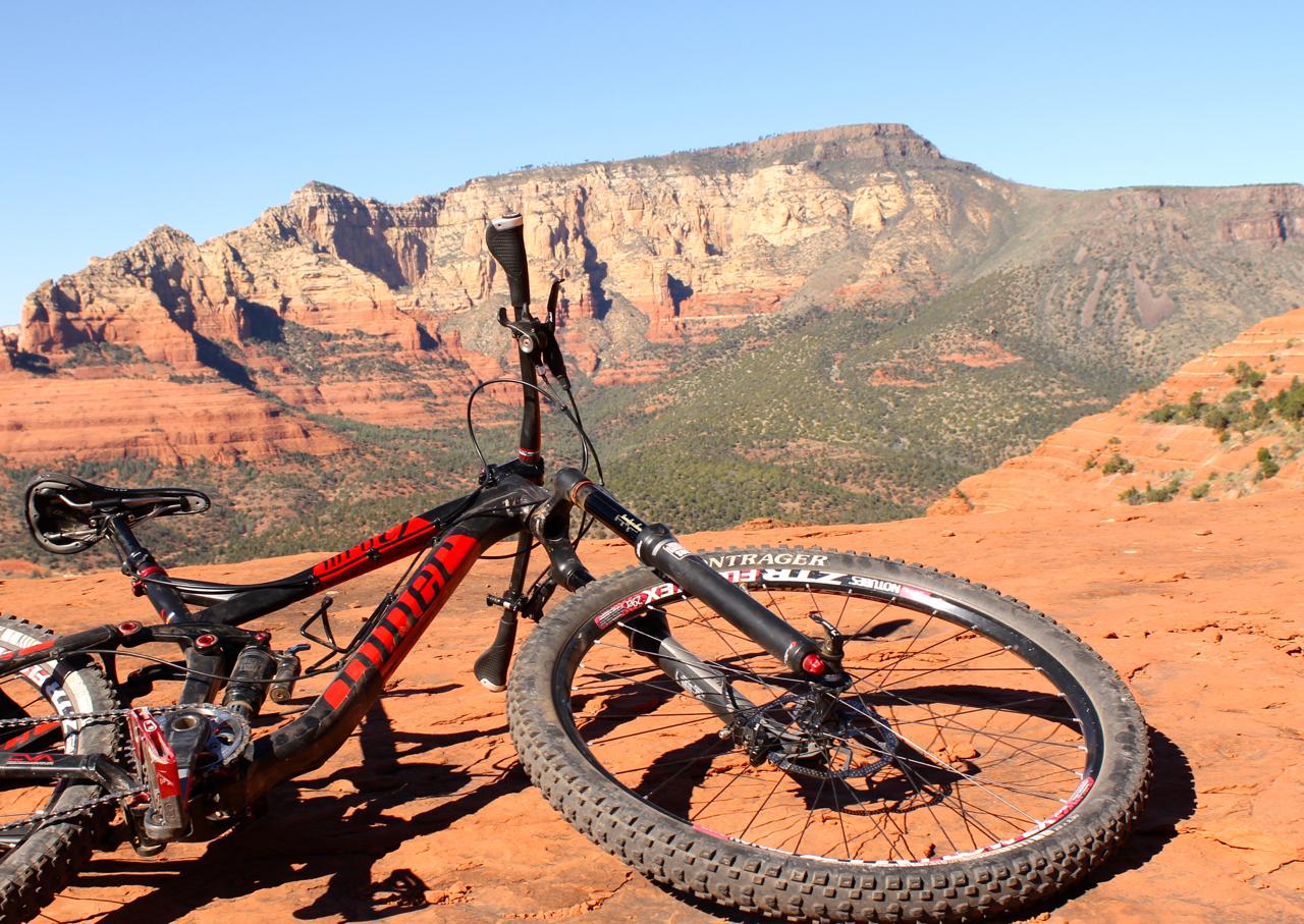 A black and red mountain bike resting on a rocky terrain with a scenic view of red rock cliffs and green hills in the background under a clear blue sky. Hangover mountain bike trail.