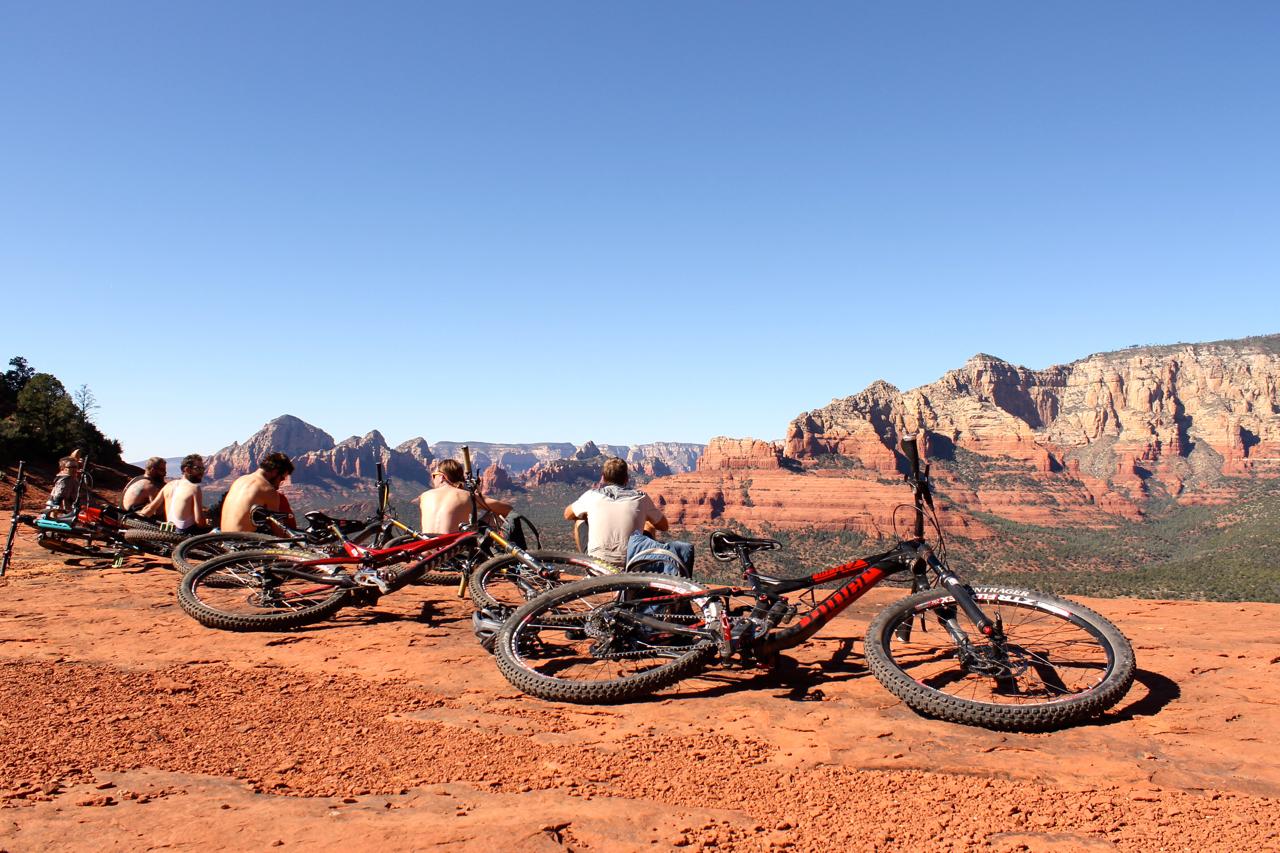 A group of people sitting on a rocky outcrop, overlooking a panoramic view of red rock formations and distant mountains. Several mountain bikes lay on the ground nearby, and the sky is clear and blue. The individuals are casually dressed and appear to be enjoying the scenery. Hangover mountain bike trail.