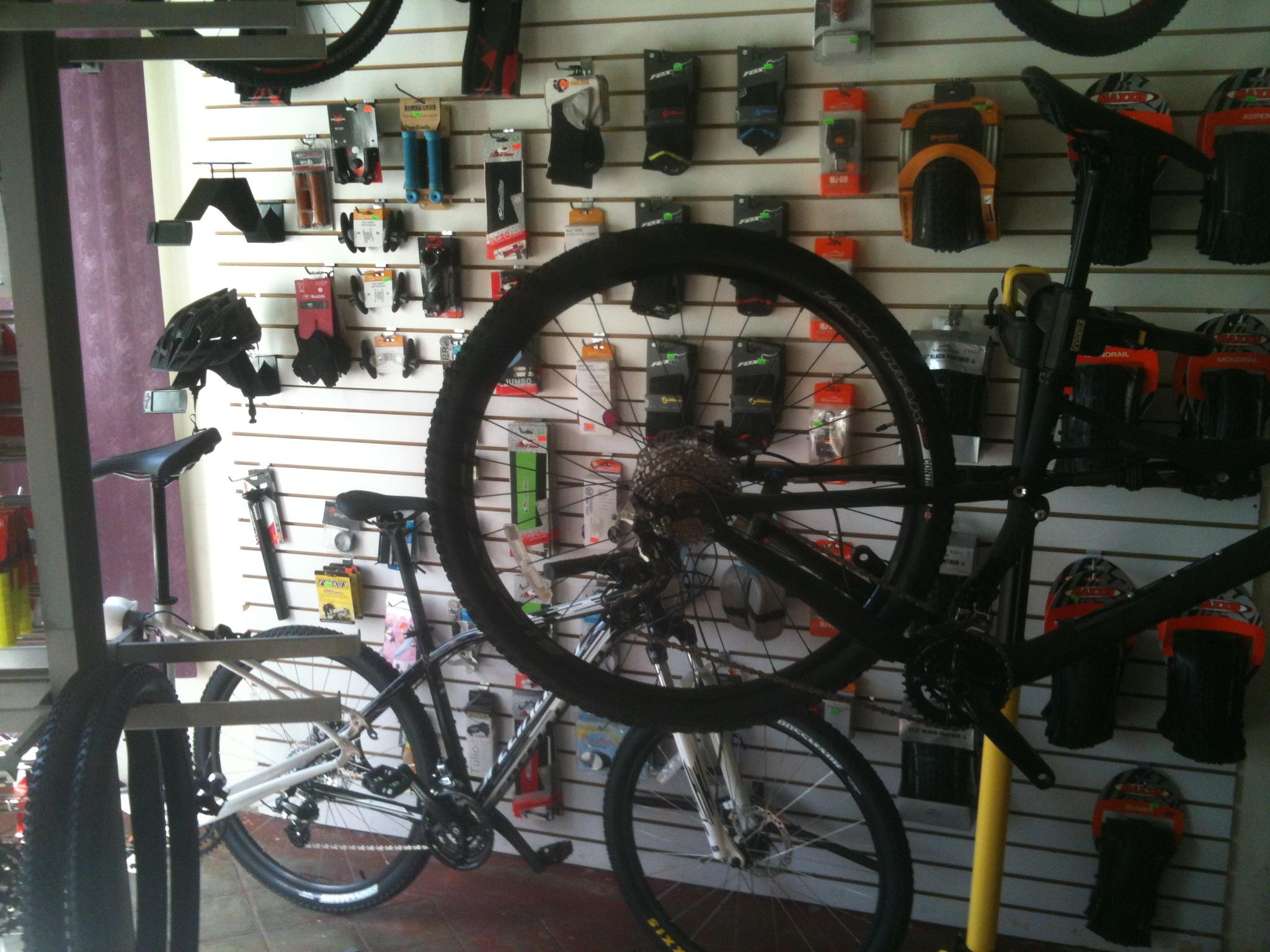 A bike shop interior featuring various bicycles and accessories displayed on slat walls. In the foreground, a black bicycle is hung up, displaying its frame and gears, while a white bicycle is partially visible behind it. The walls are lined with bike helmets, pumps, tires, and other cycling gear.