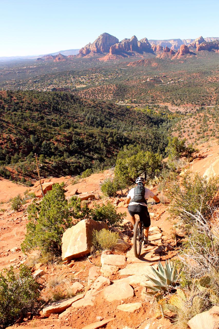 A mountain biker navigates a rocky trail in a rugged desert landscape, with lush green vegetation and distant red rock formations under a clear blue sky. Hangover mountain bike trail.