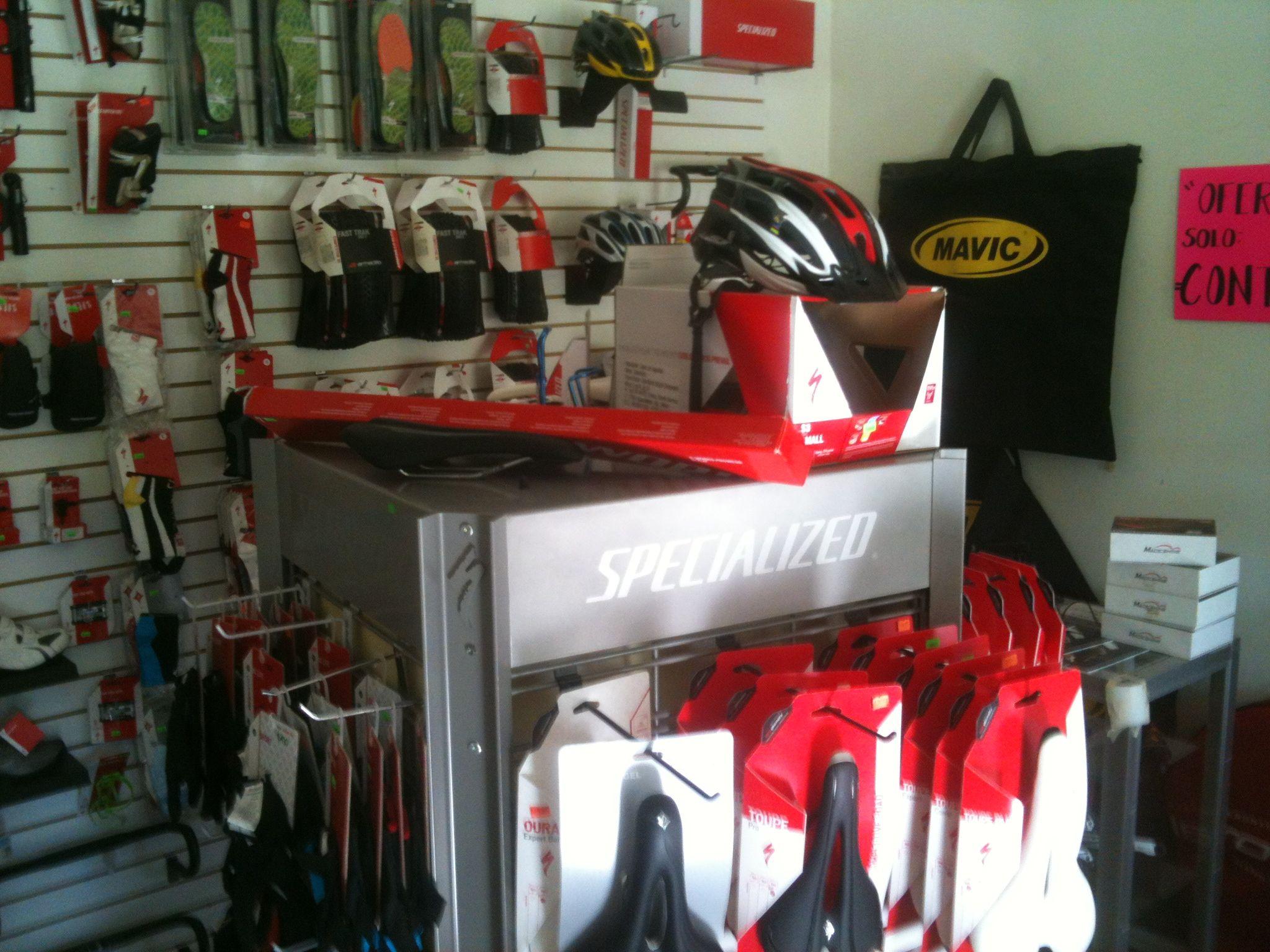 Image of a bike shop display featuring various cycling gear. The display includes a specialized table showcasing bike seats and helmets. The background is lined with hanging gloves and other cycling accessories, with a Mavic tote bag visible on the wall.