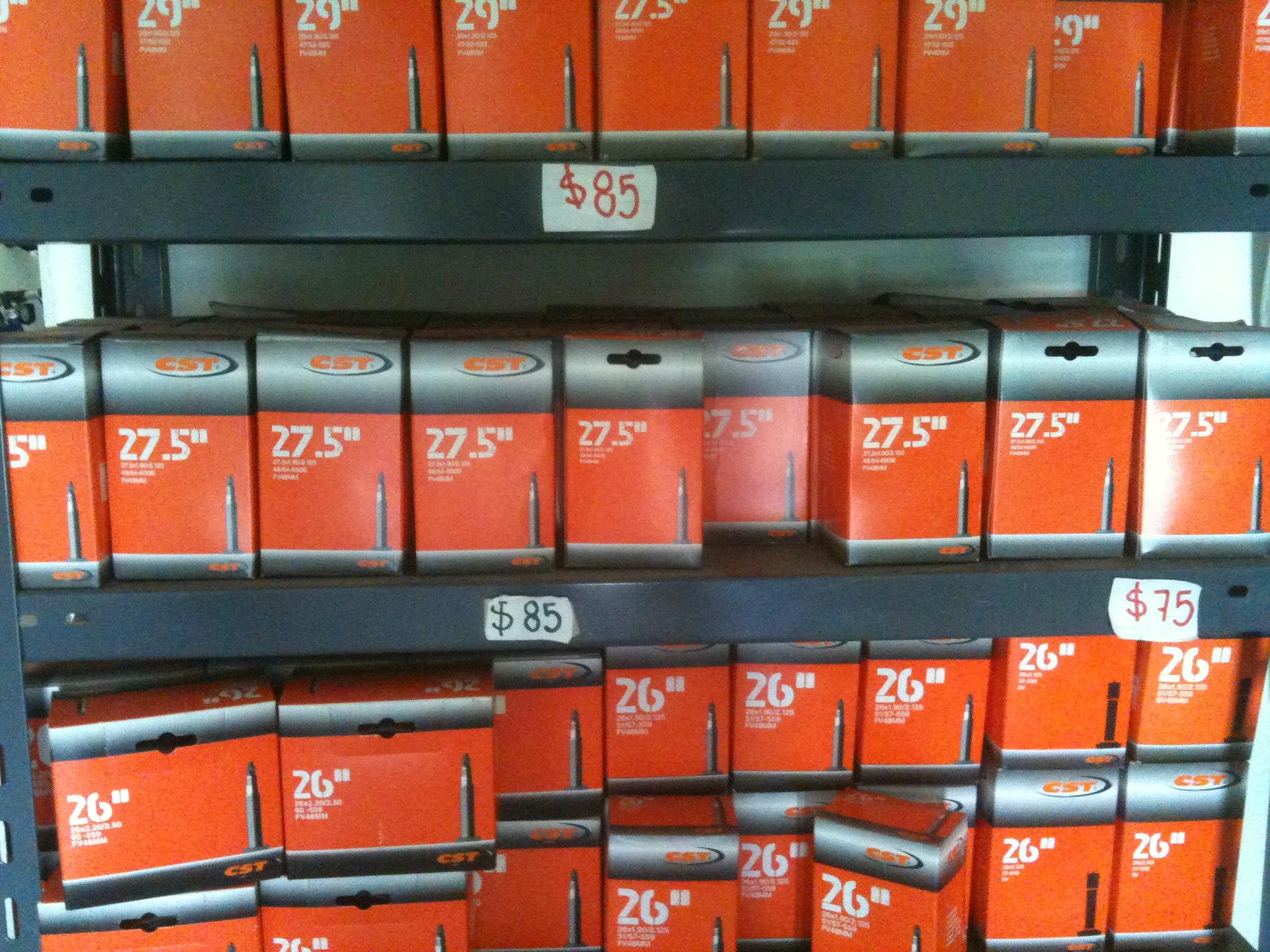 Image of a display shelf filled with boxes of CST bicycle tires. The boxes are in shades of orange and gray, labeled with different sizes (26", 27.5", and 29") and prices ranging from $75 to $85. The arrangement shows several boxes stacked in an organized manner.