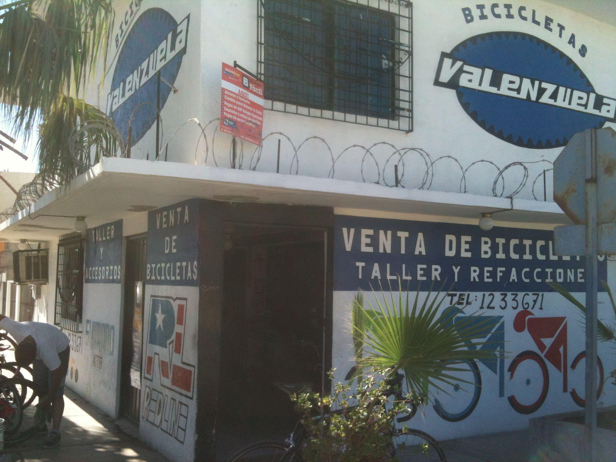 A bicycle shop with the name "Valenzuela" displayed prominently on the exterior wall, featuring signs indicating it sells bicycles and accessories. The building has a white façade with blue and red accents, and a small person is seen working on a bicycle in front of it. There is barbed wire above the windows and a palm plant in front of the shop.