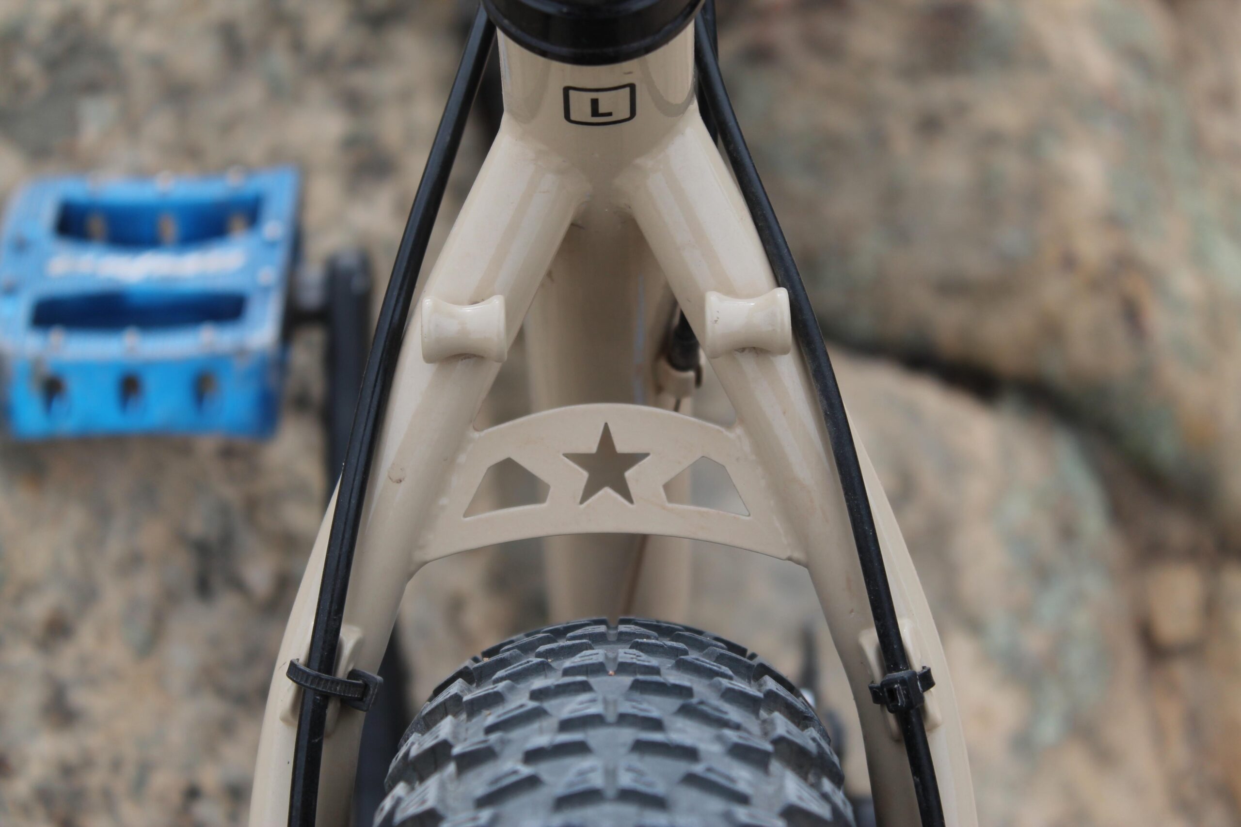 Chumba Ursa: Close-up view of a bicycle frame featuring a beige color with a star cutout design on the rear triangle. The bike's black tire is visible, highlighting its textured tread, while a blue pedal is partially in the frame's foreground against a rocky background.