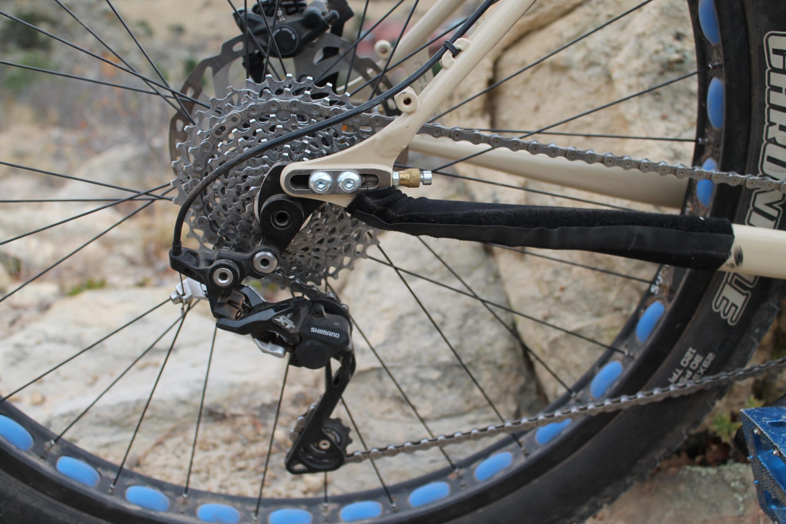 Chumba Ursa: Close-up view of a bicycle's rear derailleur and cassette, featuring a Shimano component, set against a rocky background. The image highlights the bicycle's drivetrain, including the gears, chain, and wheel with blue rim accents.