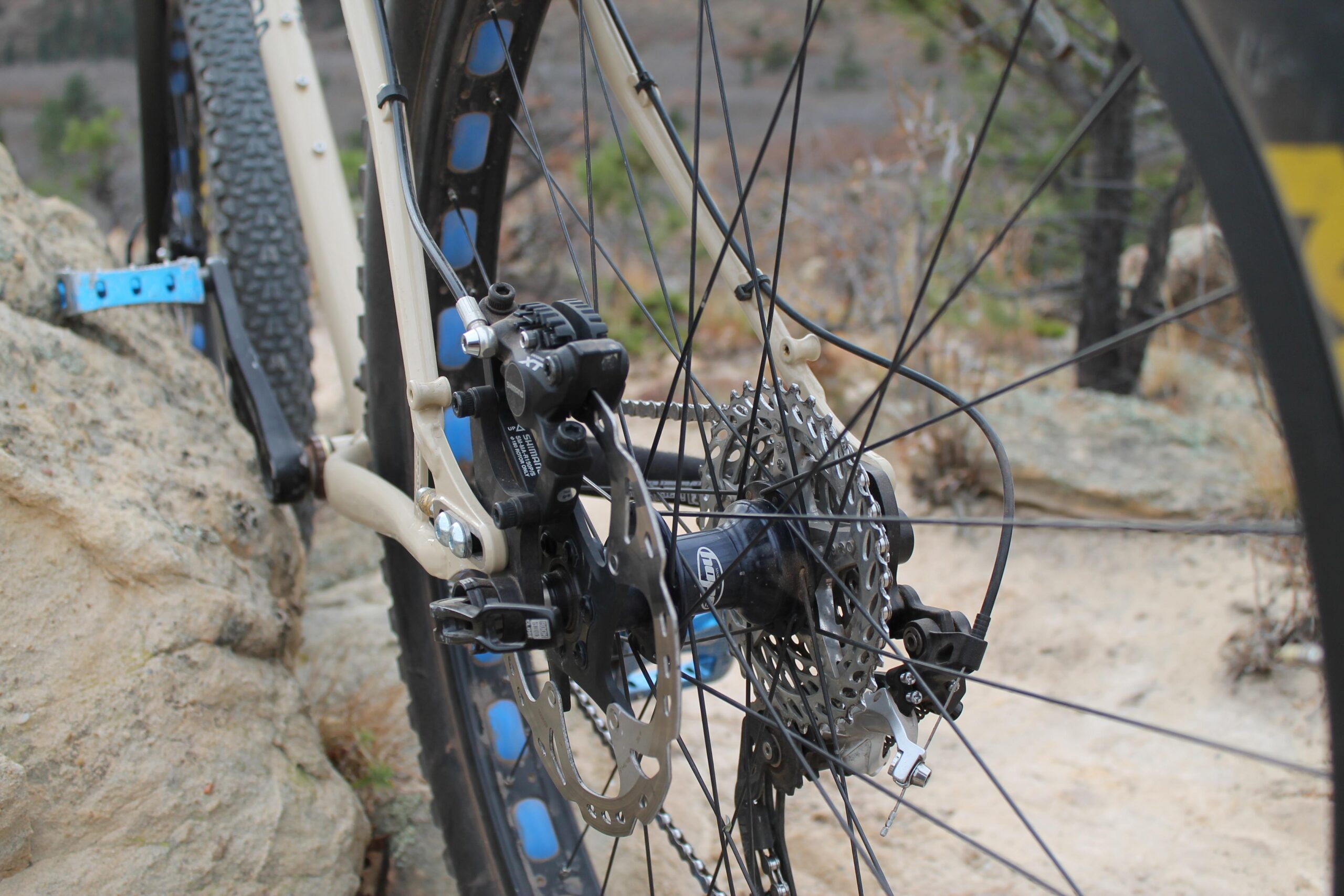 Chumba Ursa: Close-up view of a bike's rear wheel and drivetrain, showcasing the derailleur, cassette, and brake components, set against a rocky terrain with blurred greenery in the background.