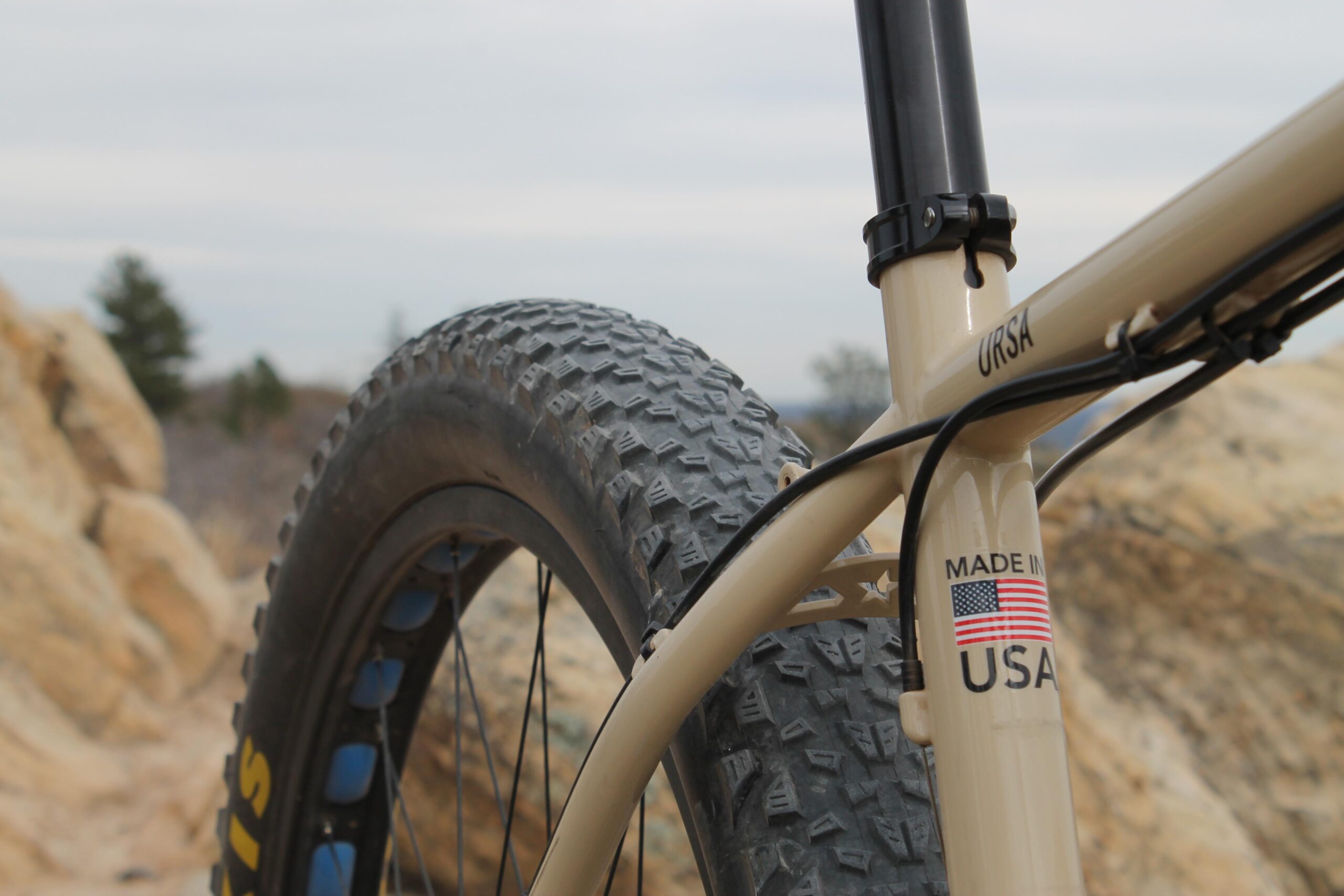 Chumba Ursa: Close-up of a mountain bike showcasing a large, textured tire and part of the bike frame. The frame is a light beige color with a label reading "MADE IN USA" featuring an American flag. The background features rocky terrain and a cloudy sky.