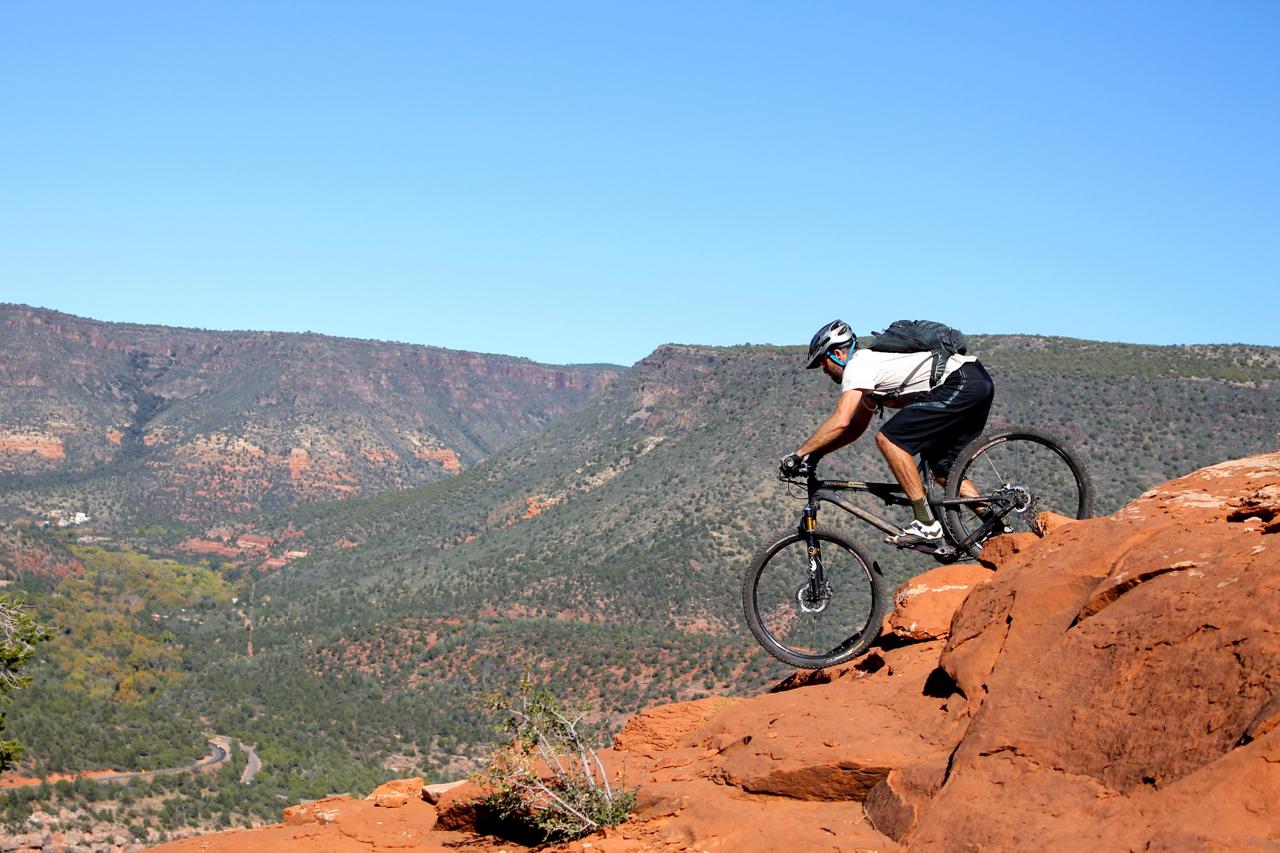 Mountain biker navigating rocky terrain on a trail, with a backdrop of expansive red rock formations and green valleys under a clear blue sky. Hangover mountain bike trail.