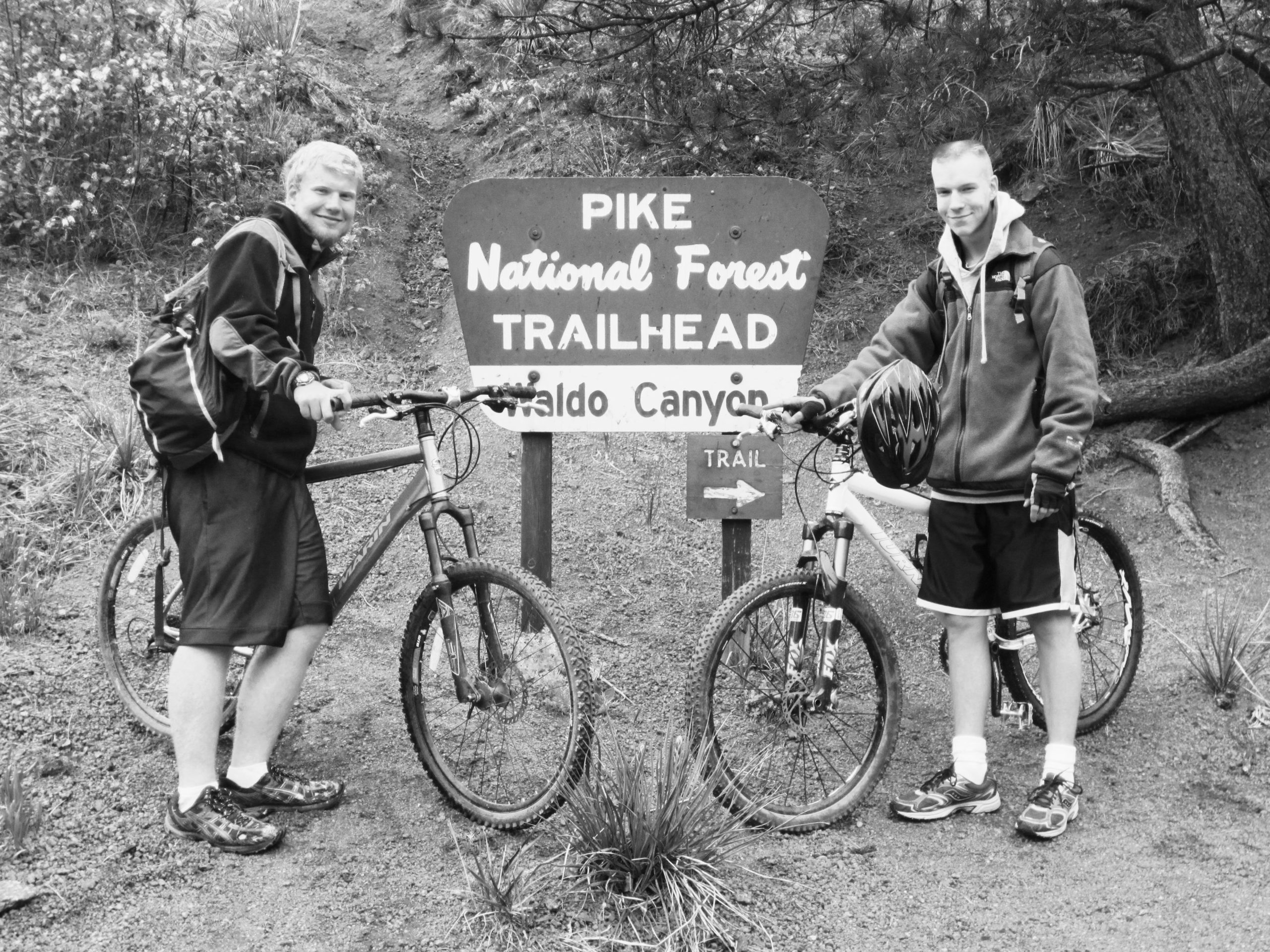 Two young men stand next to their mountain bikes at the Pike National Forest trailhead sign for Waldo Canyon. They are smiling and dressed in casual athletic wear, with one holding a bike helmet. The scene is captured in black and white, showcasing the natural surroundings and the trail ahead. Waldo Canyon mountain bike trail.