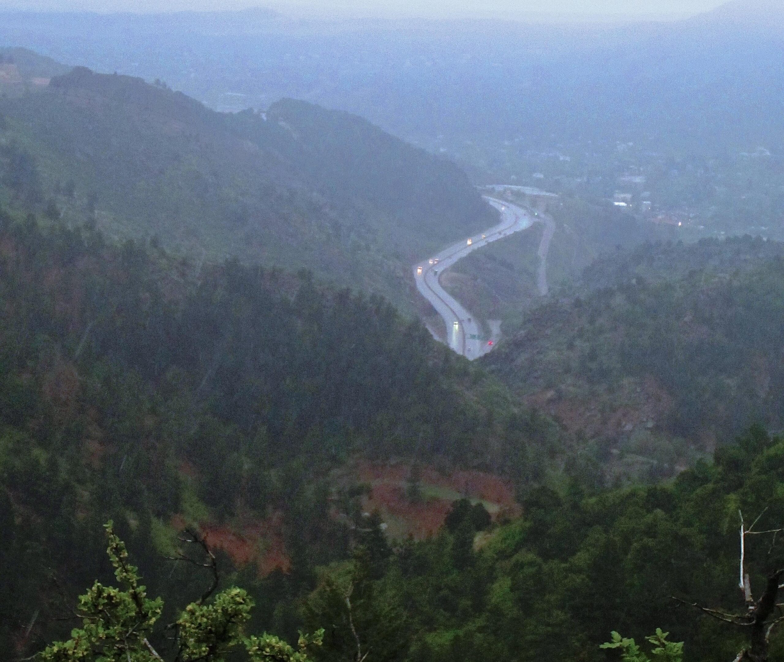 A winding road cuts through a mountainous landscape, surrounded by dense green trees and under a moody, overcast sky. The scene is captured in soft lighting, suggesting early morning or late afternoon. Small vehicles can be seen traveling along the road, with a hint of mist in the air, creating a serene and atmospheric view of nature and infrastructure. Waldo Canyon mountain bike trail.