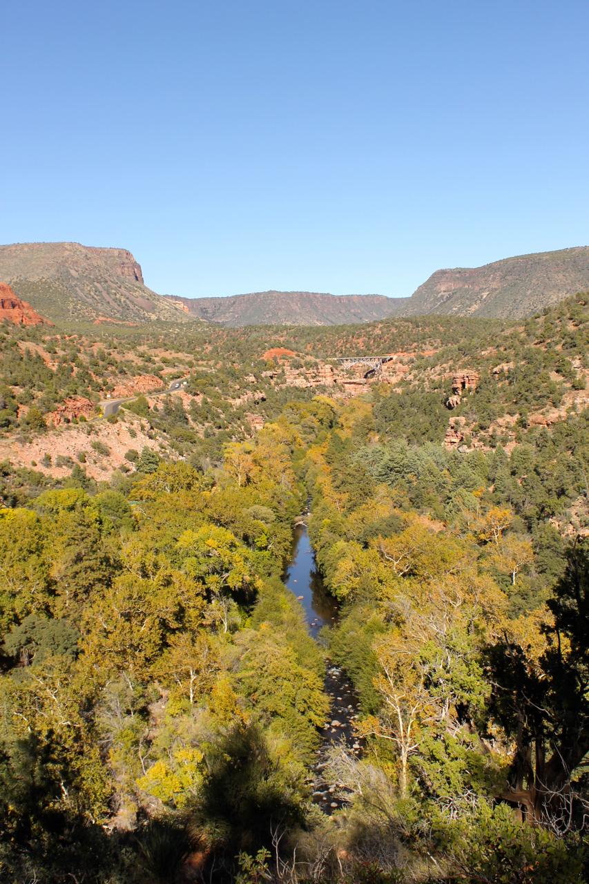 A scenic landscape featuring a winding river surrounded by lush greenery and autumn-colored trees, set against a backdrop of red rock formations and mountains under a clear blue sky. Huckaby mountain bike trail.