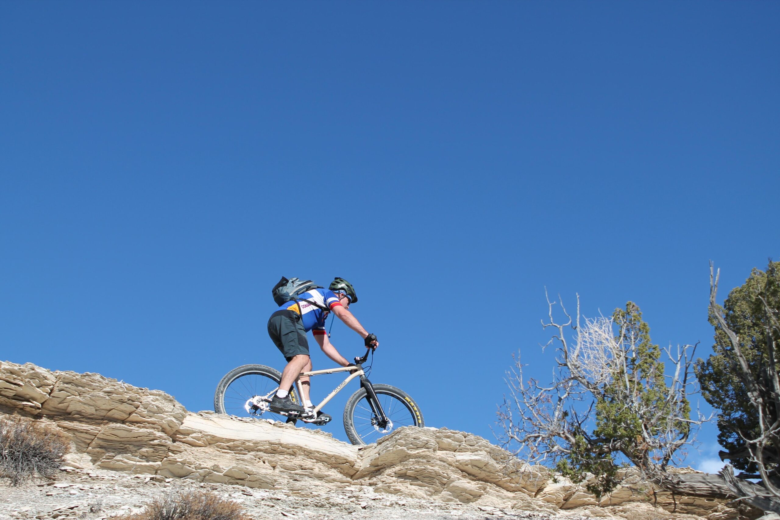 A mountain biker navigates a rocky terrain under a clear blue sky. The cyclist, wearing a helmet and a sporty outfit, appears focused as they ascend a rugged slope, showcasing a blend of adventure and athleticism. Sparse vegetation and a few trees are visible in the background, highlighting the natural landscape. North Shore Lake Pueblo State Park mountain bike trail.