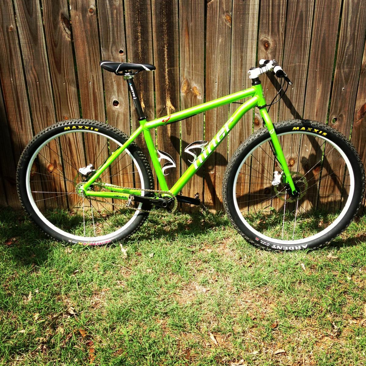 Niner One 9: A bright green mountain bike with white accents is leaning against a wooden fence. The bike features a lightweight frame, thick tires, and a single gear system, resting on grass in a sunny outdoor setting.