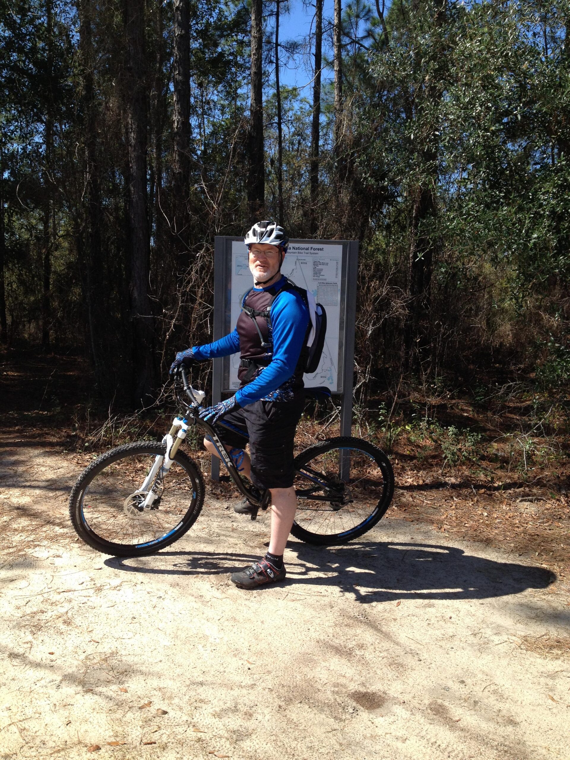 Trek Fuel EX 7 29: A mountain biker wearing a helmet and cycling gear stands next to a bike on a dirt path, with a map board in the background surrounded by trees. The scene is set in a sunny, wooded area, showcasing a recreational outdoor atmosphere.