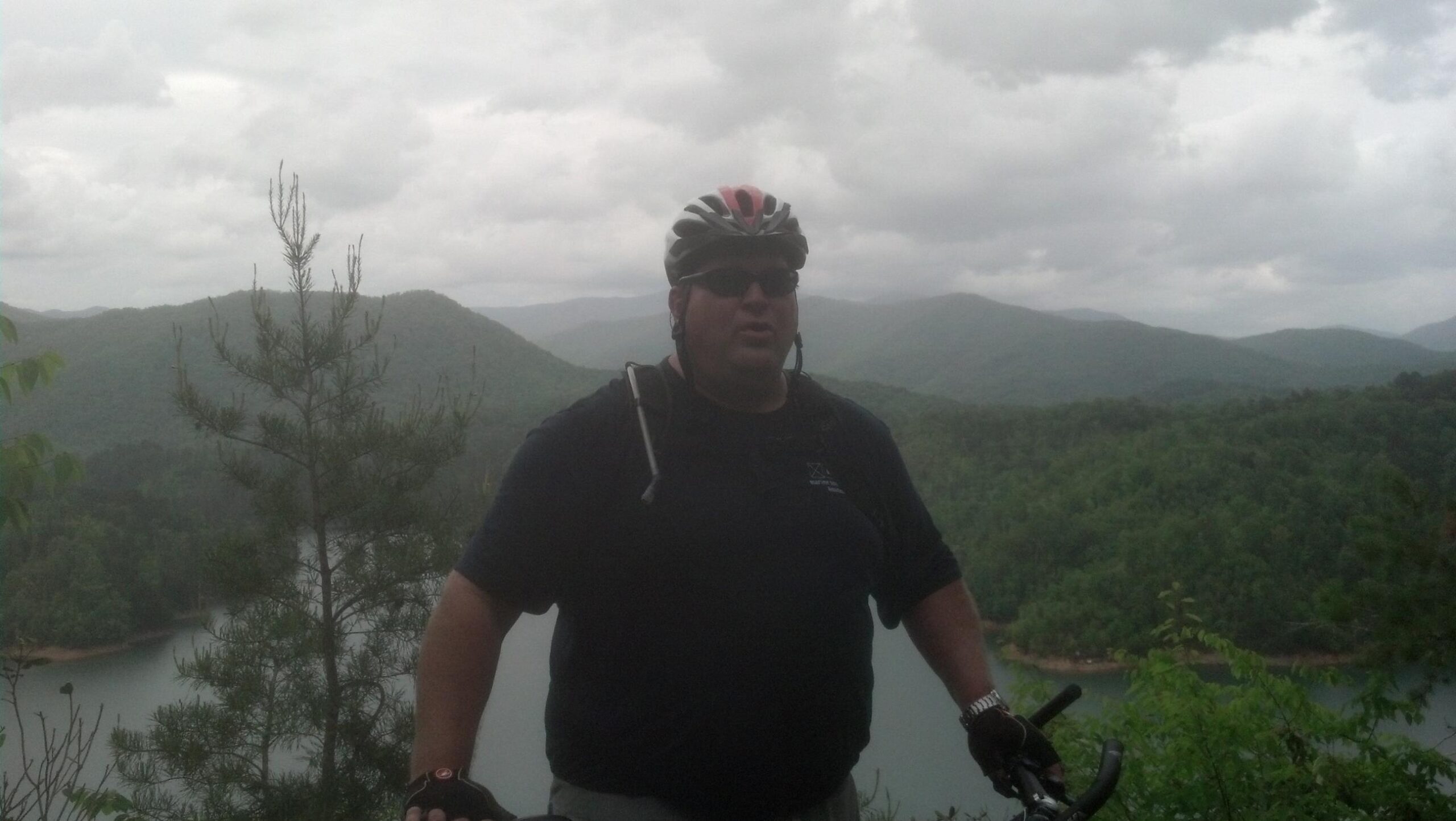 A man wearing a cycling helmet and sunglasses stands on a hillside overlooking a scenic landscape of mountains and a lake, with overcast skies in the background. He is wearing a black shirt and cycling gloves, and there are trees in the foreground. Tsali Left Loop mountain bike trail.