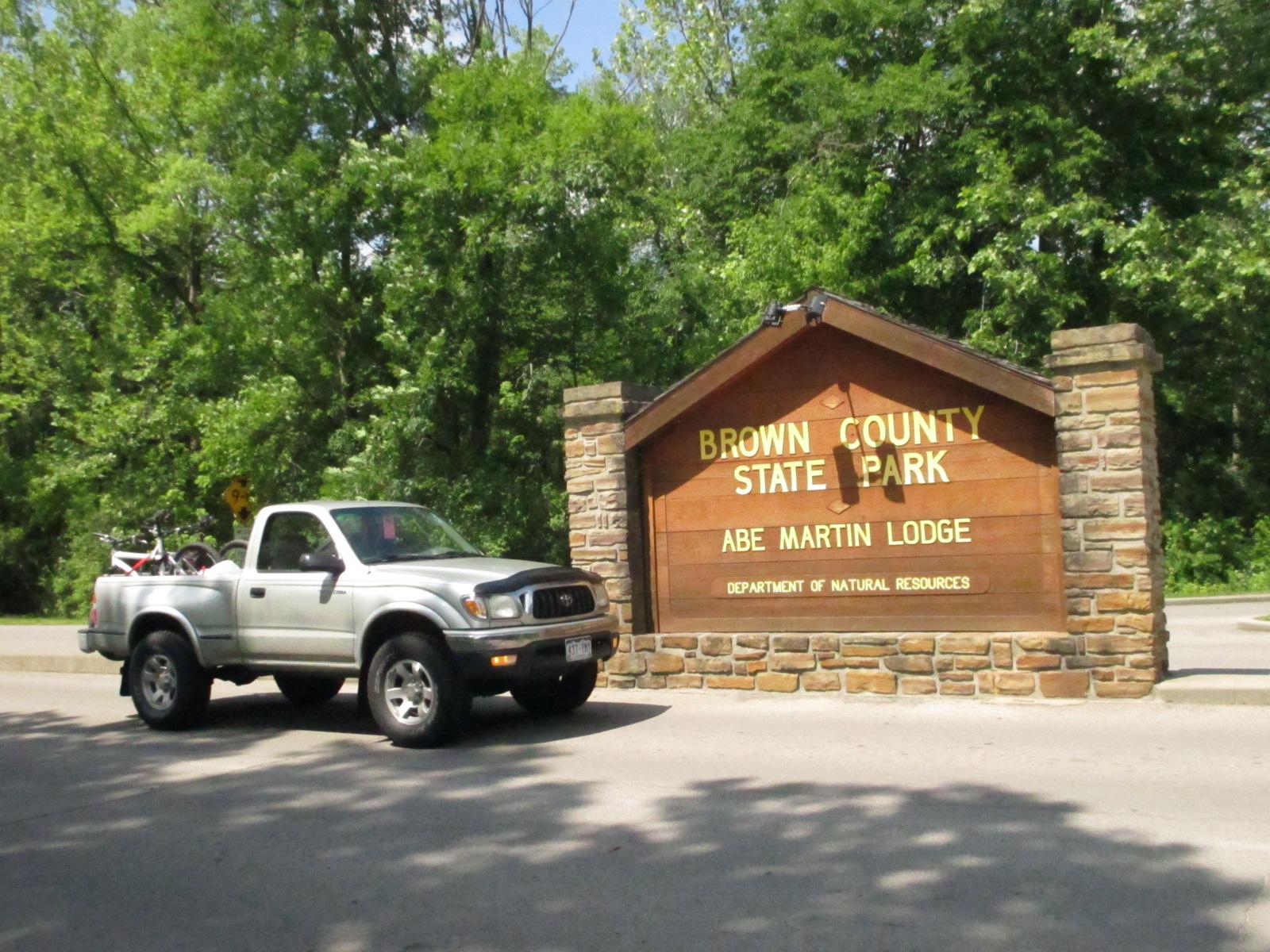 A silver pickup truck parked near the entrance sign of Brown County State Park, which displays "BROWN COUNTY STATE PARK" and "ABE MARTIN LODGE" in bold yellow lettering. The background features lush green trees under a clear sky. Brown County Park mountain bike trail.