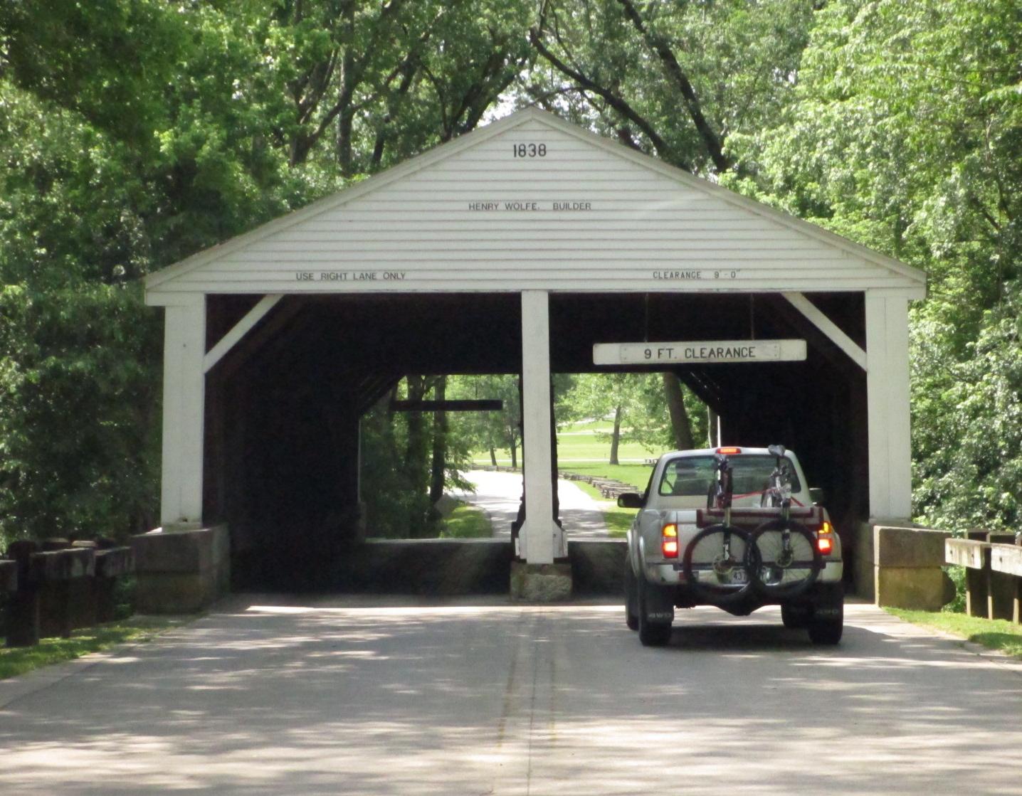 A covered bridge from 1838 with a vehicle approaching, showing a sign for "9 ft. clearance" and "Use right lane only." The bridge is surrounded by lush greenery, and a bicycle rack is visible on the back of the vehicle. Brown County Park mountain bike trail.