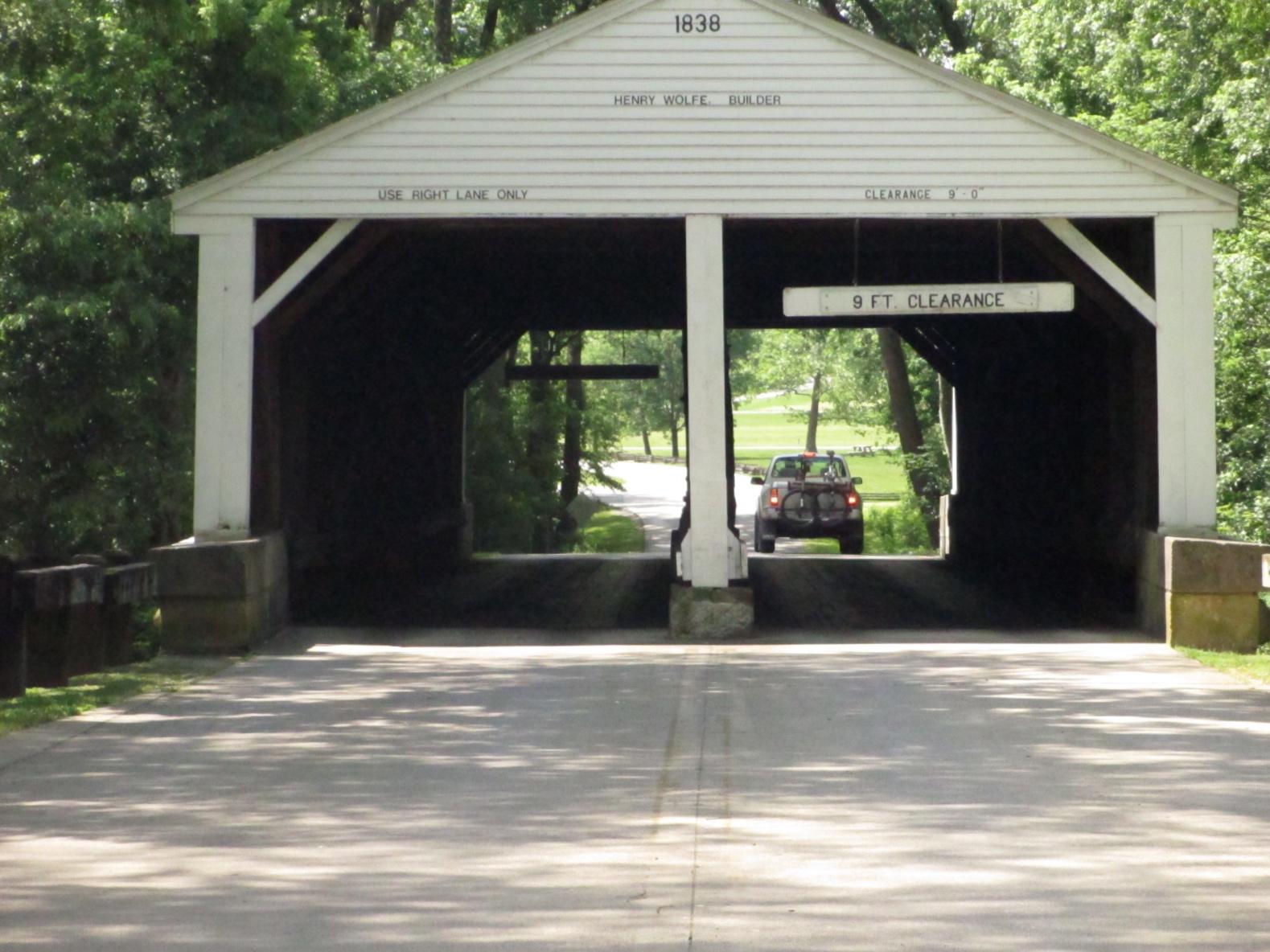 A covered bridge with a white wooden structure, featuring a sign that reads "Use Right Lane Only" and "9 ft. Clearance." A vehicle is seen approaching from inside the bridge, surrounded by greenery on both sides and a clear view of the road ahead. Brown County Park mountain bike trail.