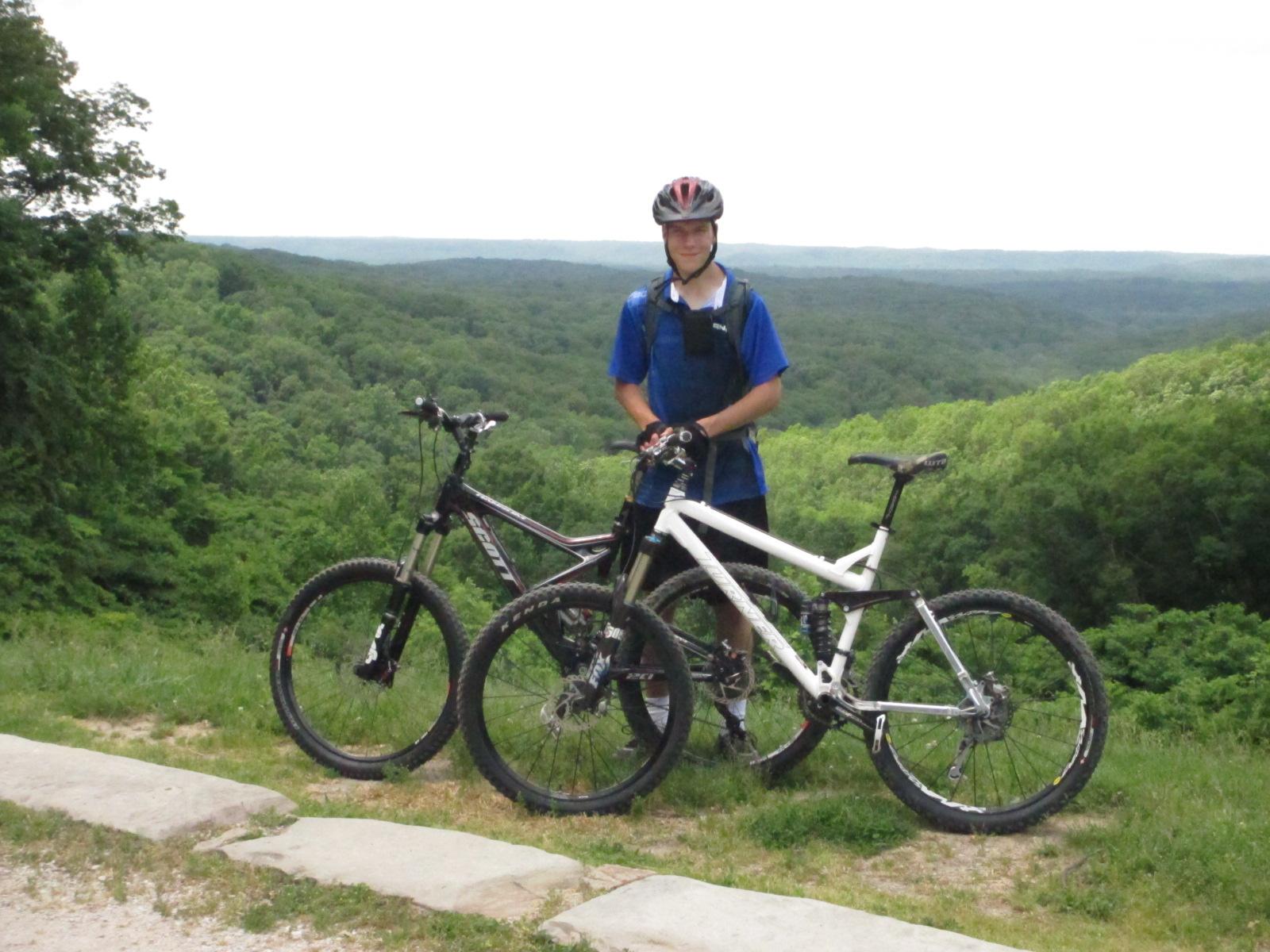 A person in a blue shirt and helmet stands next to two mountain bikes on a scenic hillside, with lush green trees and rolling hills in the background. Brown County Park mountain bike trail.