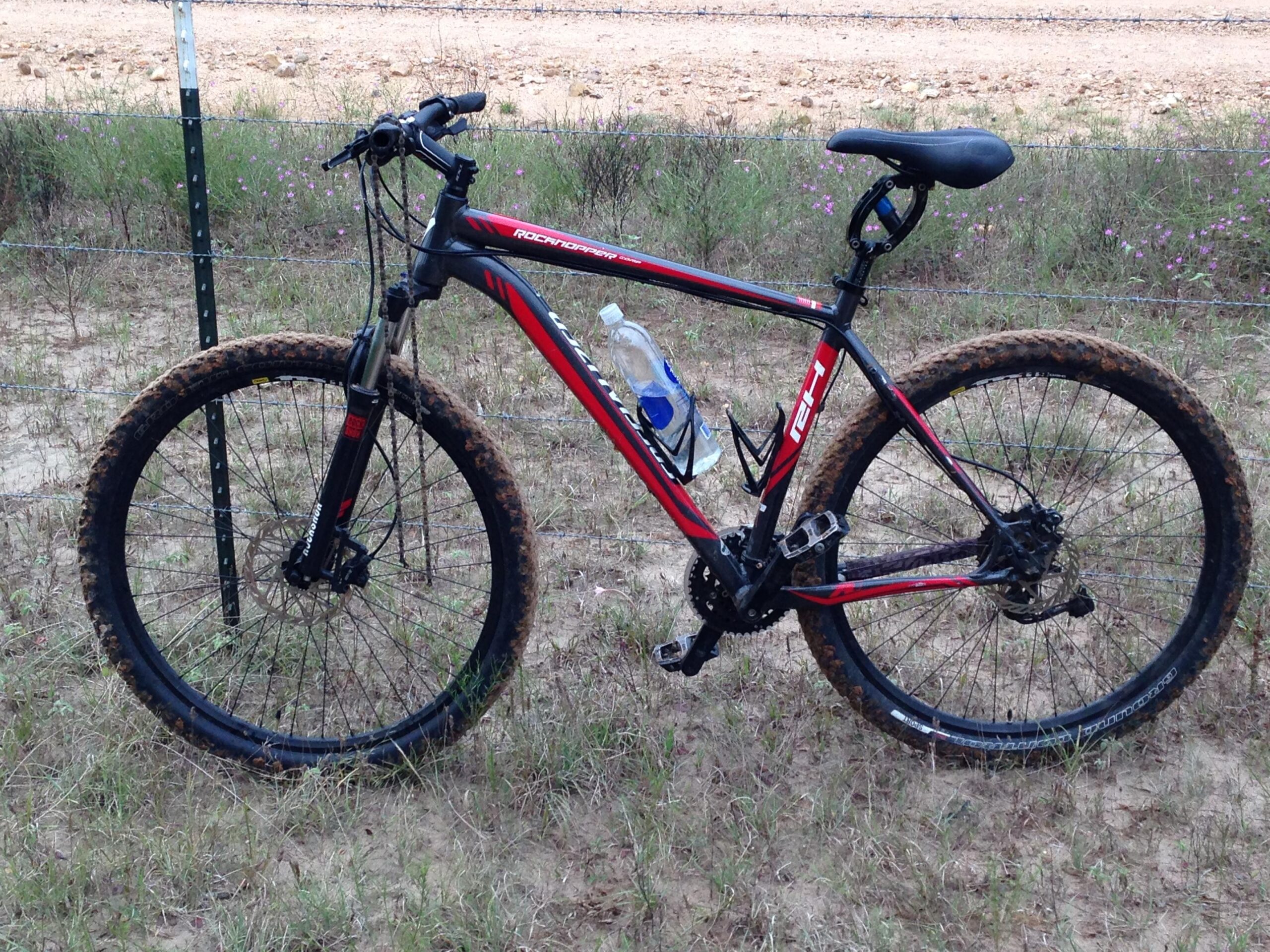 Specialized Rockhopper: A red and black mountain bike stands on a grassy area, partially obscured by dirt on the wheels. A water bottle is mounted in a cage on the frame. The background features a barbed wire fence and a gravel road.