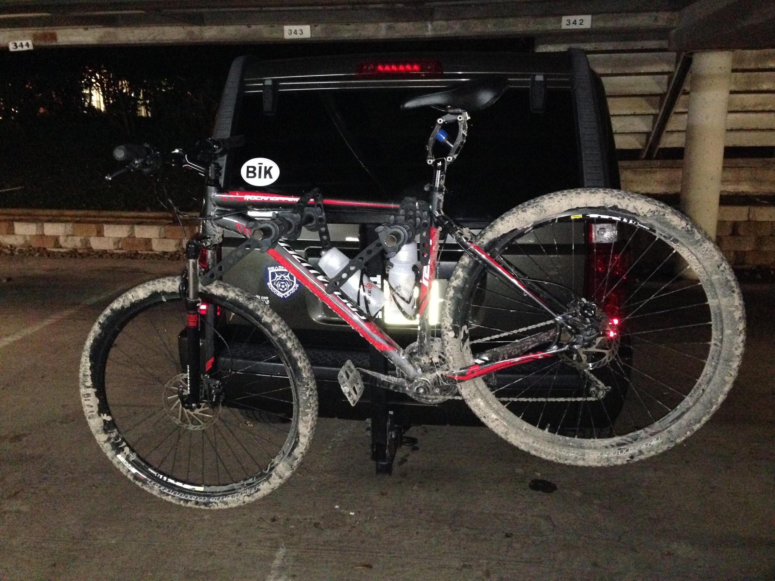 Specialized Rockhopper: A mountain bike mounted on the back of a vehicle, with visible dirt on the tires and frame, parked in a dimly lit garage. The vehicle features a sticker that reads "BIk."