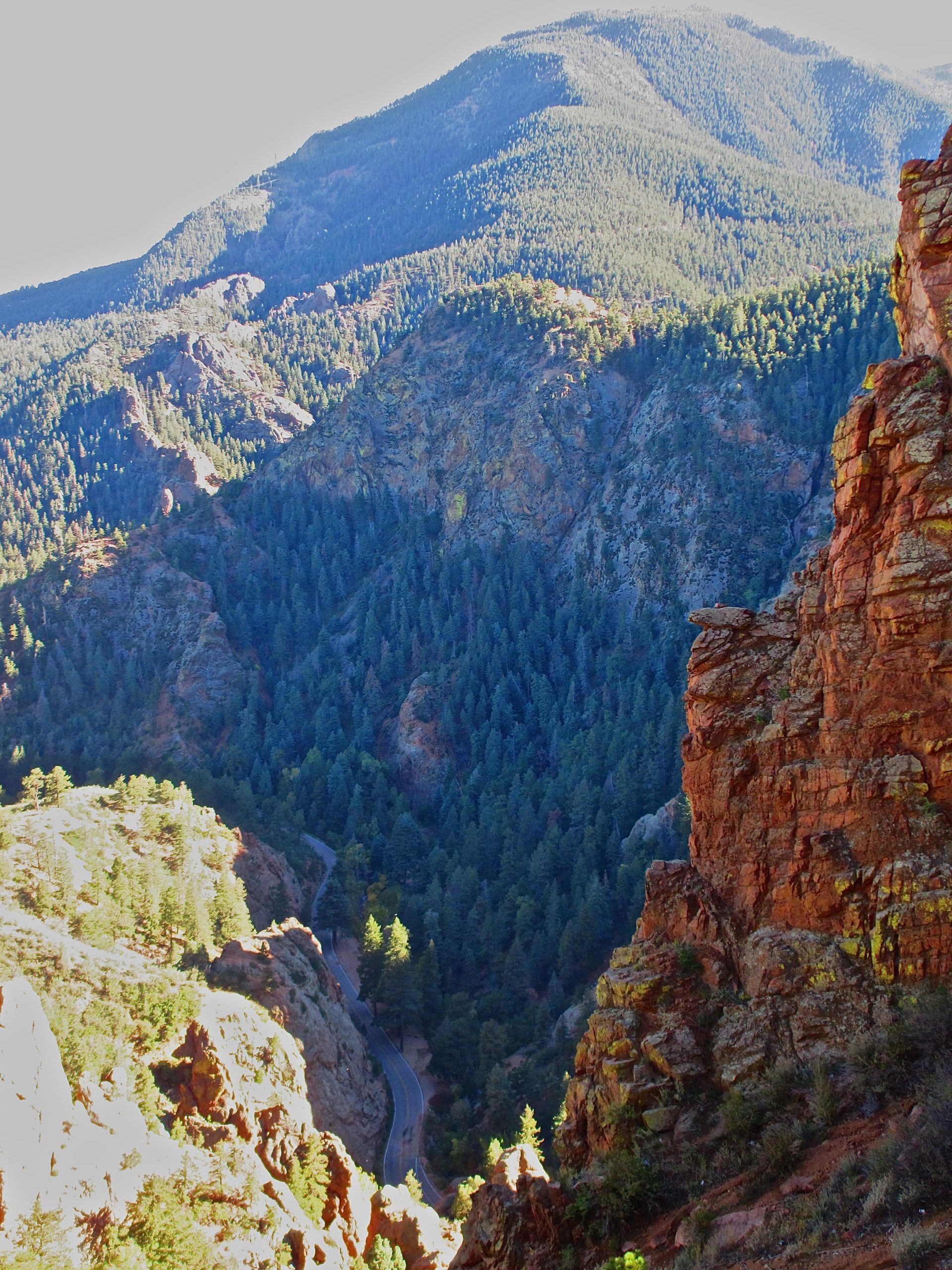 A panoramic view of a mountainous landscape, featuring steep, rocky cliffs in the foreground and lush, green pine forests in the valley. A winding road can be seen traversing through the trees, leading deeper into the scenic mountains under a clear sky. Captain Jack's mountain bike trail.
