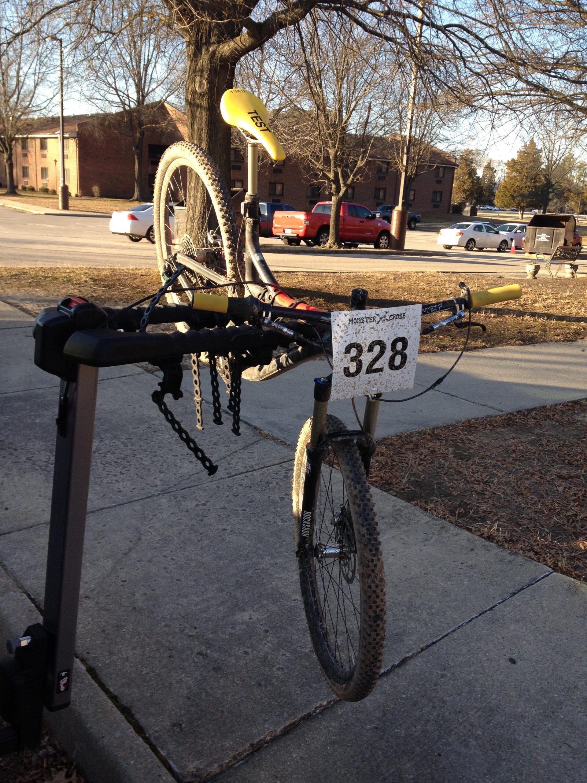 Devinci Atlas Carbon XP: A mountain bike is mounted on a bike rack with its front wheel lifted off the ground. The bike features a bright yellow saddle labeled "TEST" and a race number tag displaying "328." In the background, a tree is visible along with several parked cars and buildings. The ground is covered with leaves, indicating it's autumn or early spring.