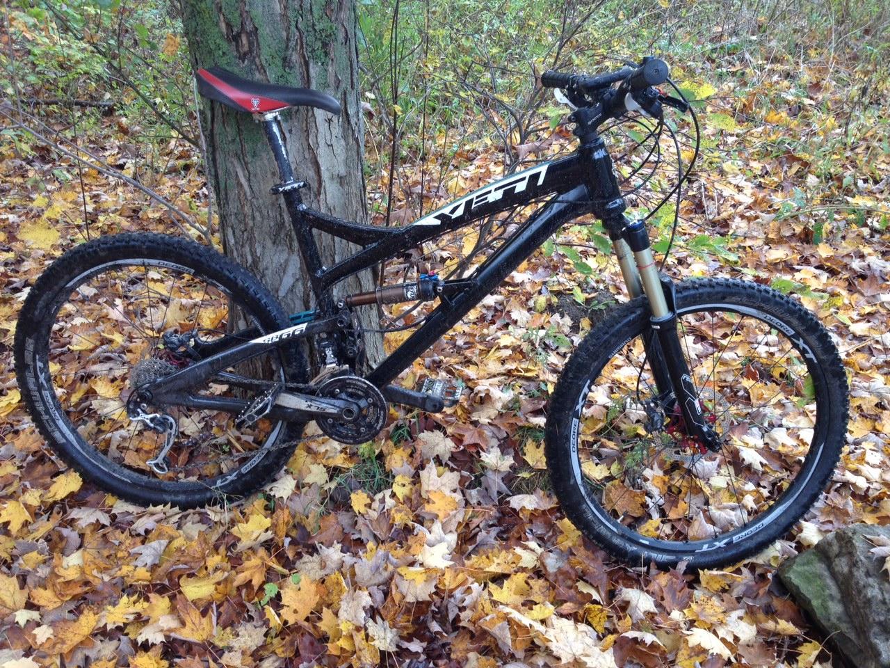 Yeti SB66: A mountain bike resting against a tree, surrounded by colorful autumn leaves. The bike features a sleek black frame, front suspension, and rugged tires, highlighting its off-road capabilities.