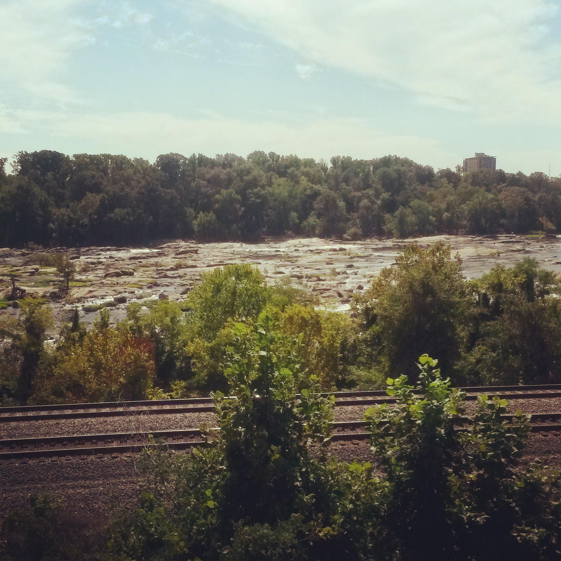 A scenic view of a rocky landscape along a river bank, framed by lush trees and greenery. In the foreground, railway tracks run parallel to the edge of the water, and the sky is partly cloudy. The rocky area is mostly dry, showcasing various shapes and sizes of rocks, with foliage adding a vibrant touch to the natural setting. Northbank Trail mountain bike trail.