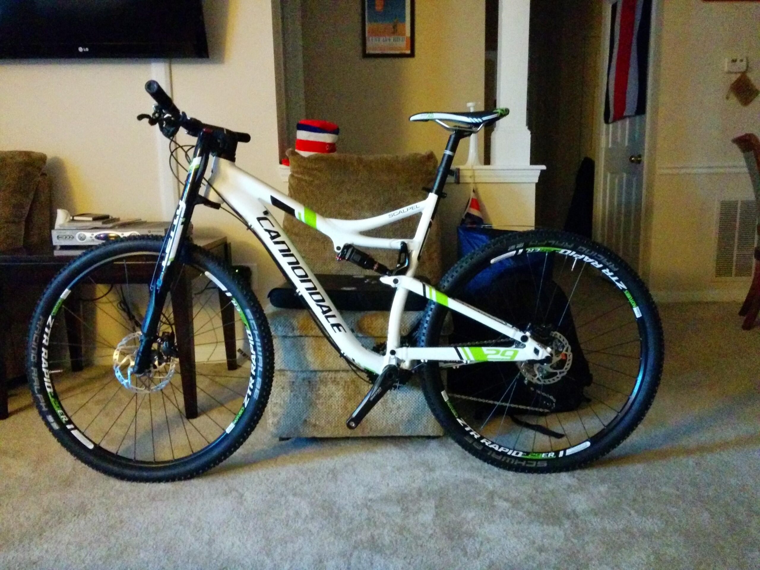 A white and green Cannondale mountain bike is positioned on a carpeted floor, leaning against a couch. The bike features thick tires and a modern design, with the seat at a height suitable for riding. In the background, there is a television and some household items, creating a casual indoor setting.