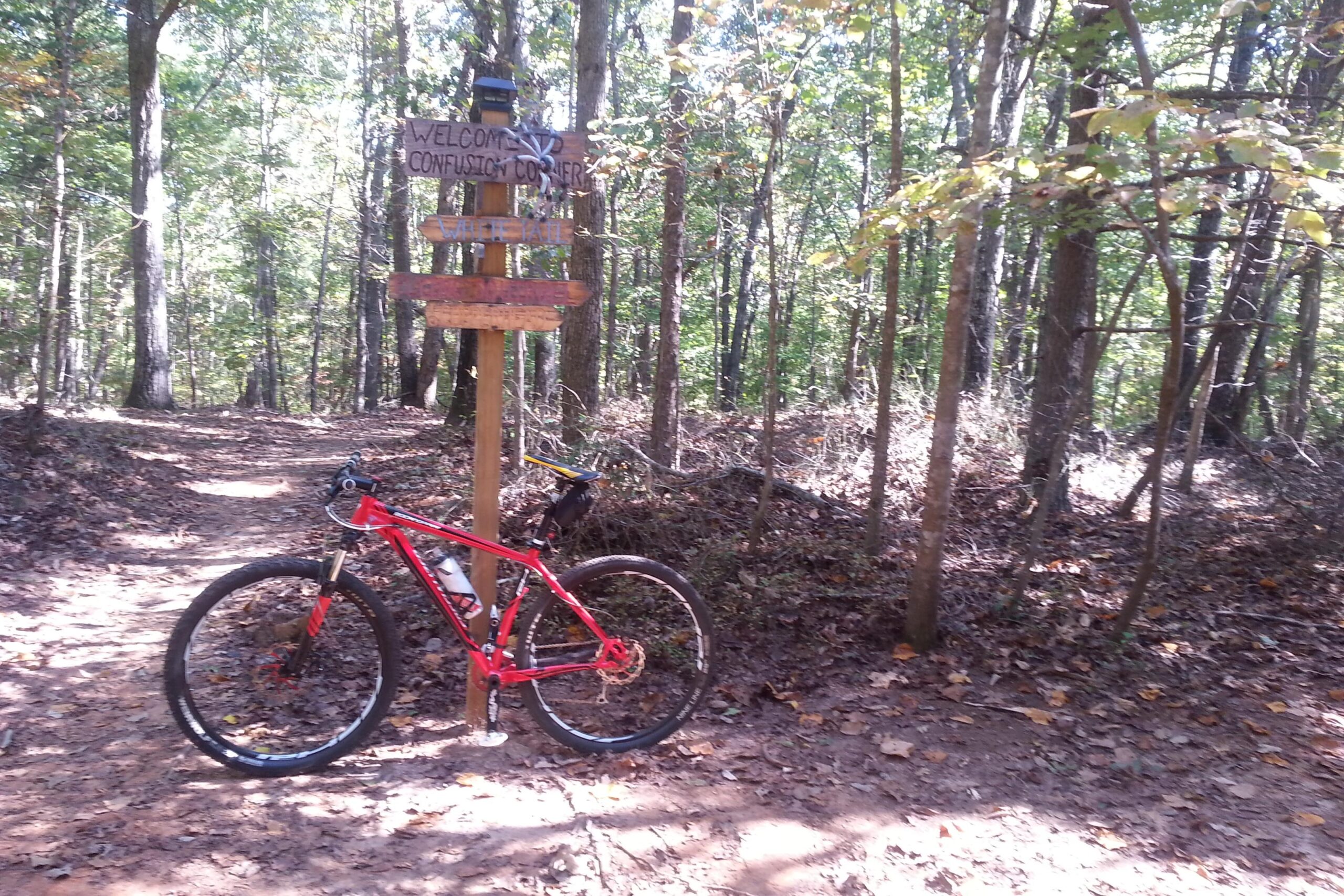 A red mountain bike is resting beside a wooden signpost that reads "Welcome to Confusion Corner" along a dirt trail in a wooded area. The trail is surrounded by tall trees, and the ground is covered with fallen leaves, indicating an autumn setting. Chicopee Woods mountain bike trail.