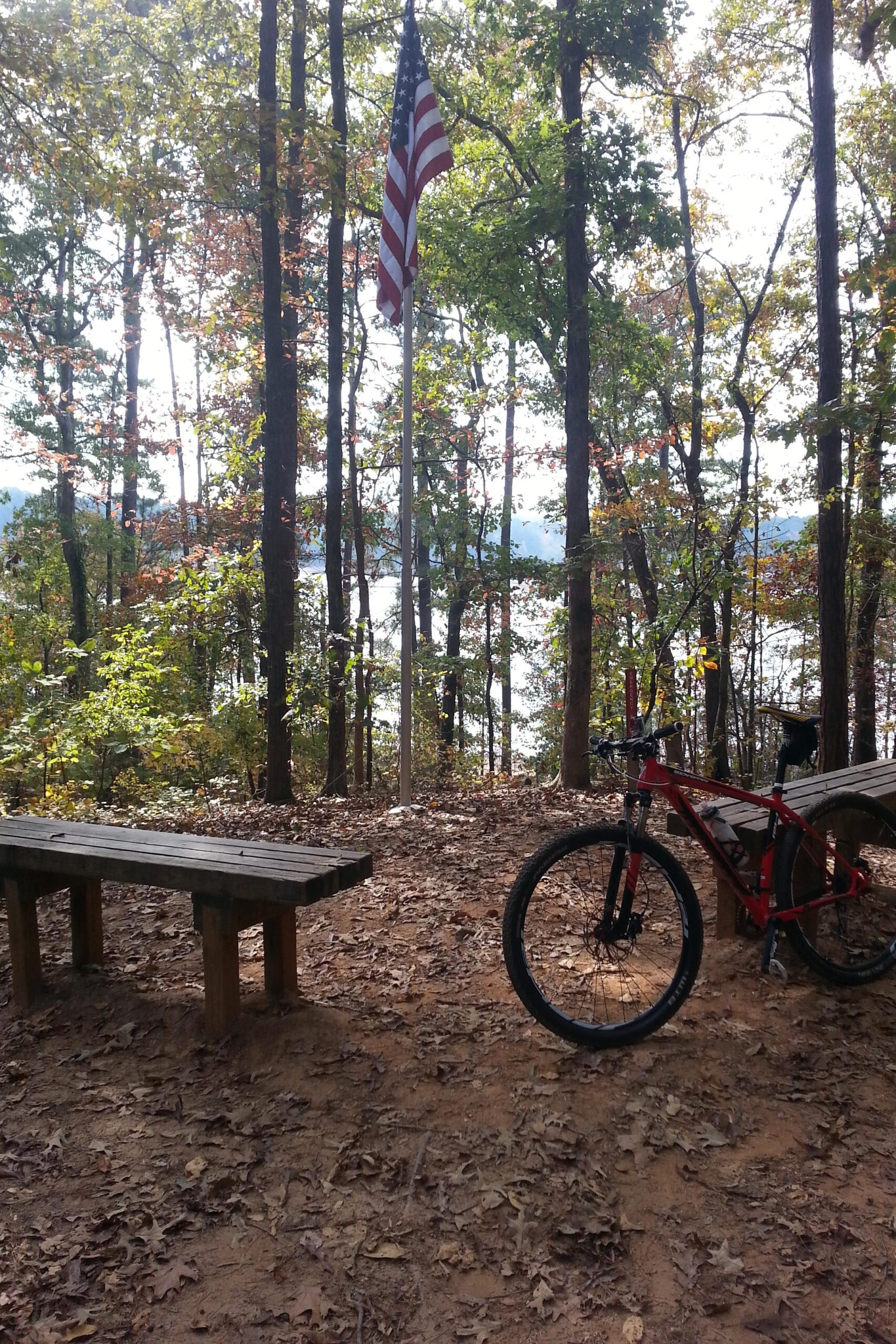 A peaceful outdoor scene featuring a U.S. flag standing tall among tall trees in a wooded area. In the foreground, there are two wooden benches, while a red mountain bike leans against one of them. The ground is covered with fallen leaves, and a glimmering lake can be seen in the background, bathed in natural sunlight. Blankets Creek mountain bike trail.