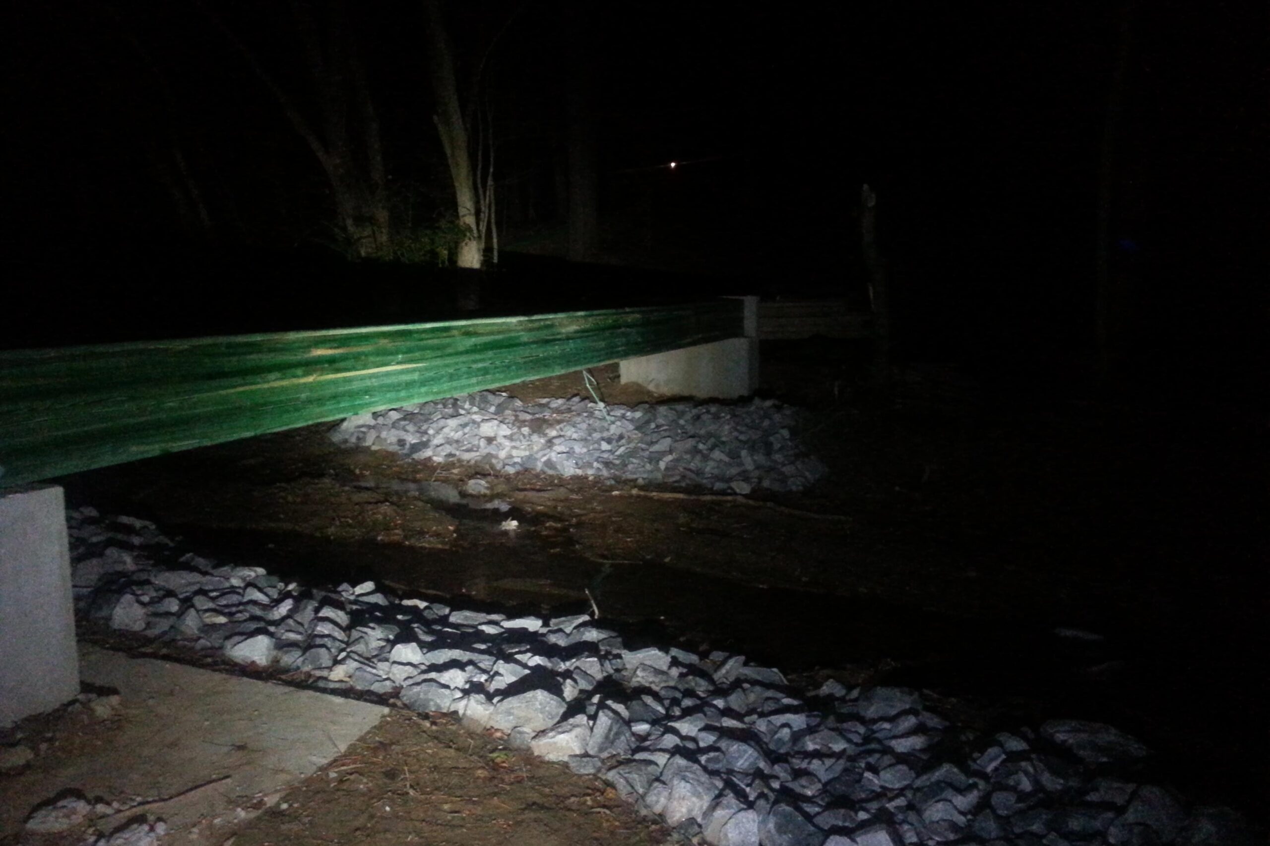 A wooden bridge with a green railing spans a small stream at night, surrounded by rocks and trees. The scene is dimly lit, with the focus on the bridge and the rocky bank below. Charleston Park mountain bike trail.