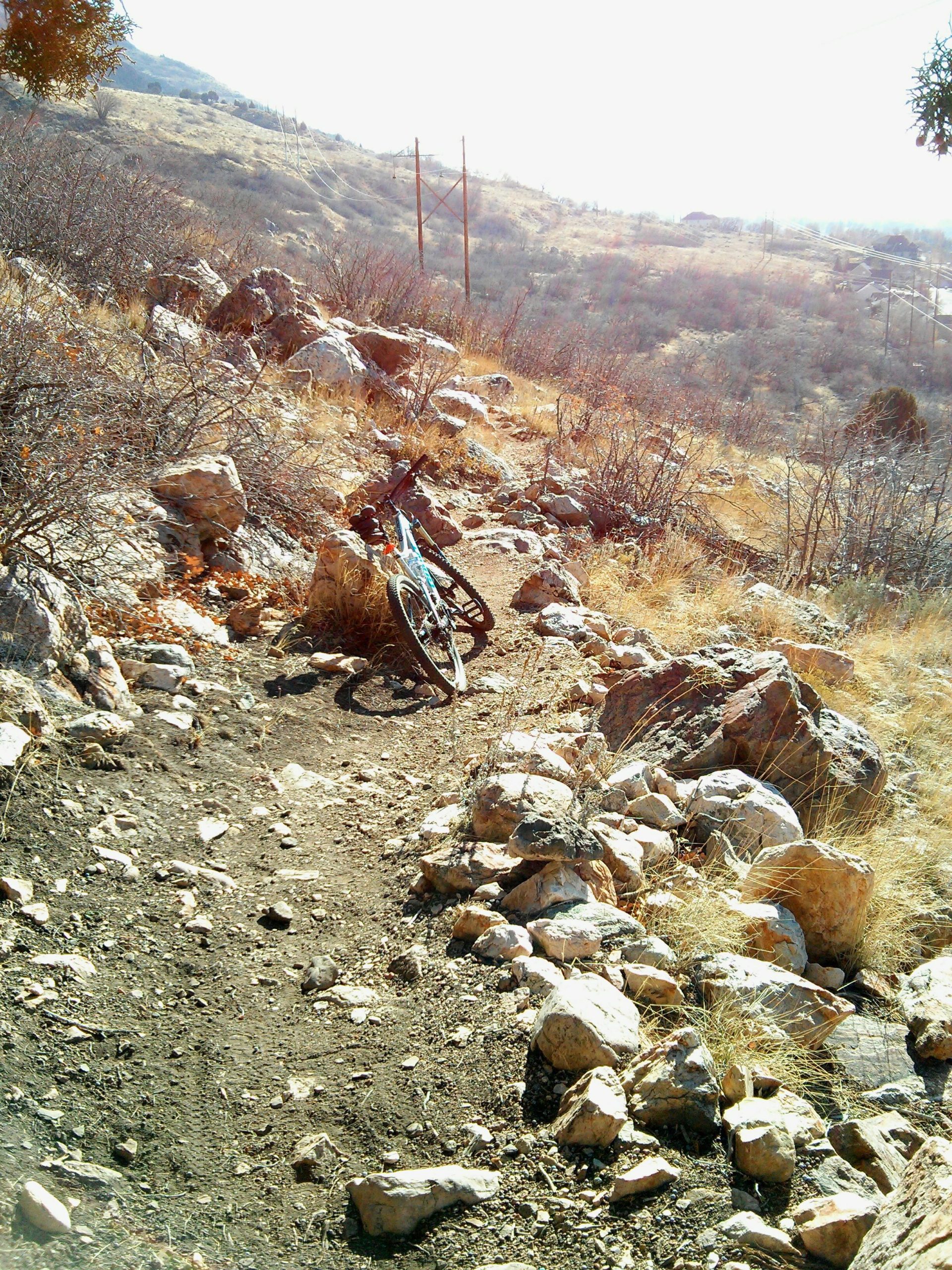 A mountain bike leaning against a rock formation on a rugged, dirt trail surrounded by dry grass and shrubs. In the background, rolling hills and power lines are visible under a bright sun. Bonneville Shoreline Ogden North Of 12th mountain bike trail.