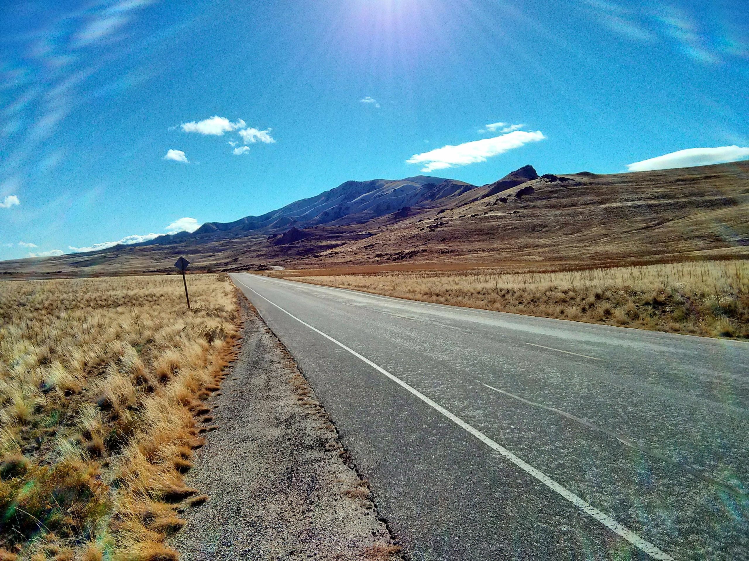 A wide, empty road stretches into the distance, flanked by golden grasslands and rocky hills under a bright blue sky with a few scattered clouds. Sunlight creates a lens flare effect, adding warmth to the serene landscape. Antelope Island mountain bike trail.