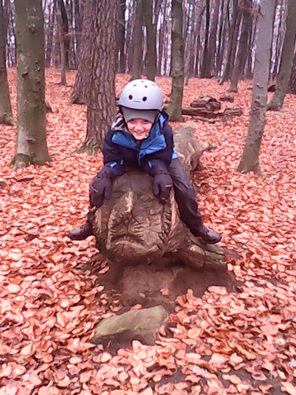 A young boy wearing a bike helmet sits on a carved wooden turtle in a wooded area covered with fallen leaves. The boy is smiling, dressed in a blue and black jacket, with trees in the background. Landstuhl Trail mountain bike trail.