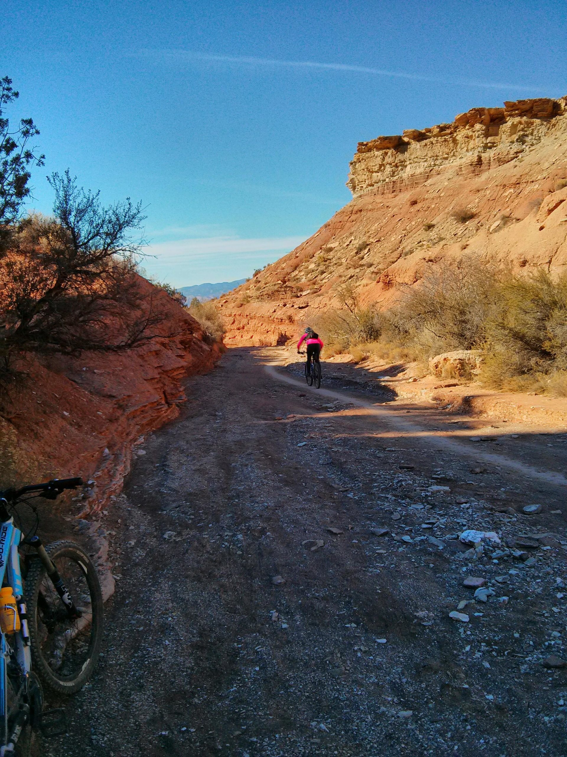 Mountain biker riding on a dirt trail surrounded by rocky terrain and shrubs, with bright blue skies overhead and red-hued cliffs in the background. A mountain bike is resting on the left side of the image. J.E.M. Trail mountain bike trail.