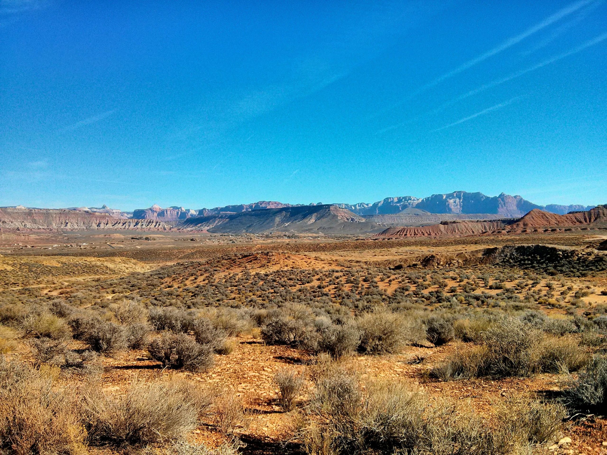 A scenic view of a vast desert landscape under a clear blue sky, featuring rolling hills, sparse vegetation, and distant mountains with unique geological formations. J.E.M. Trail mountain bike trail.