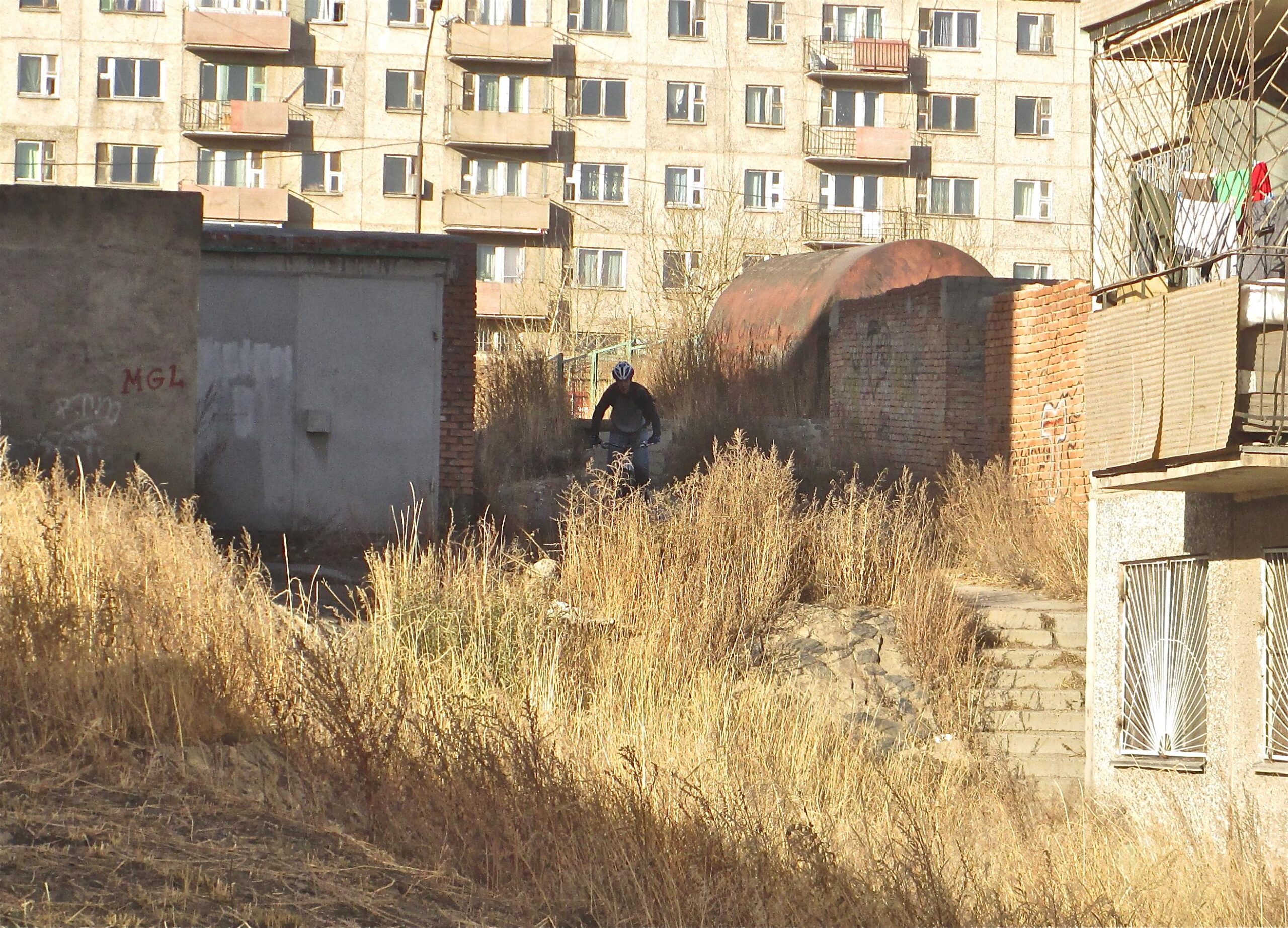 A person wearing a helmet rides a bicycle through tall, dry grass and weeds, navigating between old, partially collapsed buildings in an urban area. In the background, a multi-story apartment building with several windows and balconies is visible. The scene appears to depict a mix of urban decay and outdoor activity. District 15 Network mountain bike trail.