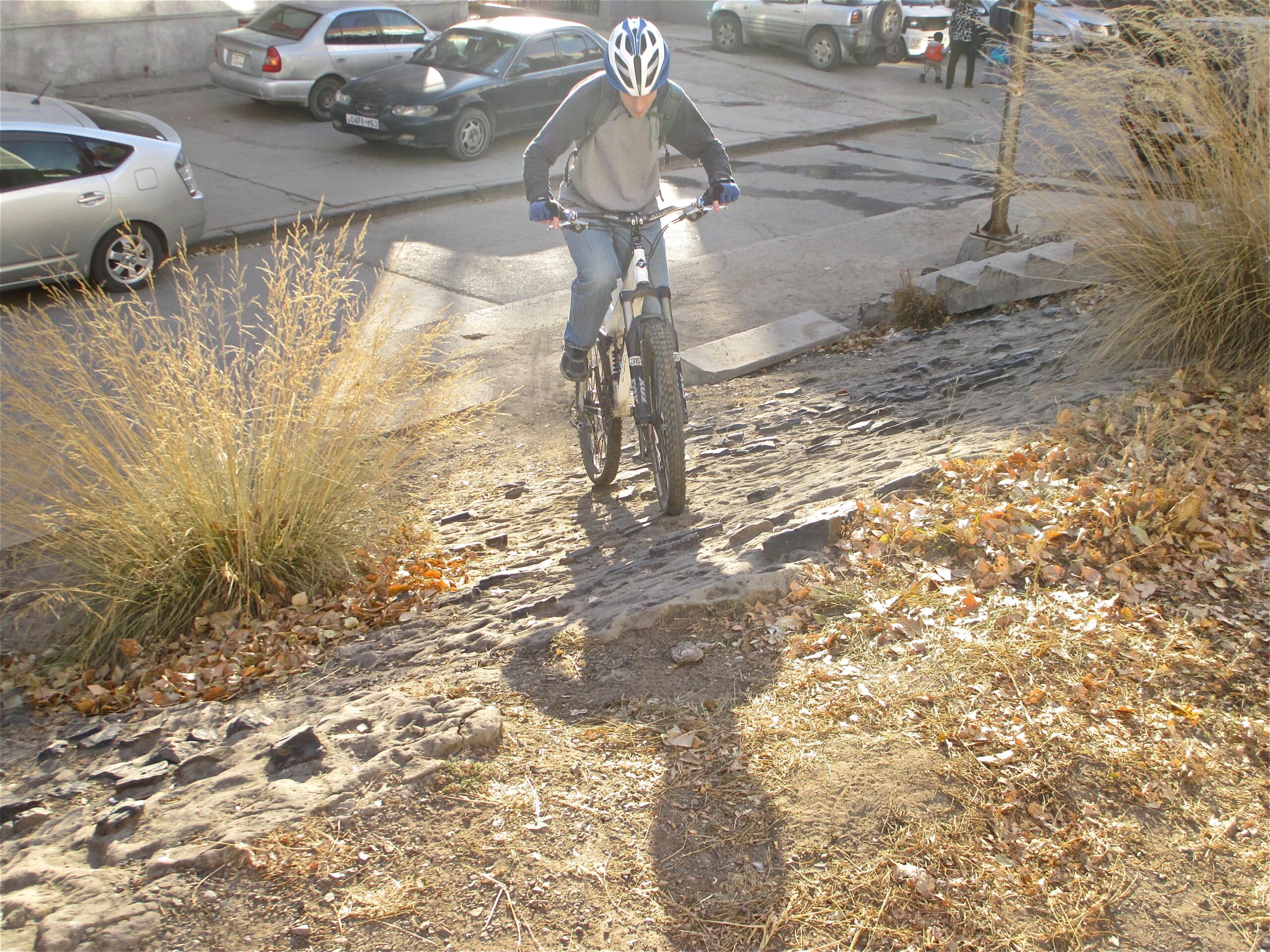 A person riding a mountain bike on a rocky path, surrounded by dry grass and fallen leaves, with parked cars in the background and a sunny atmosphere. District 15 Network mountain bike trail.