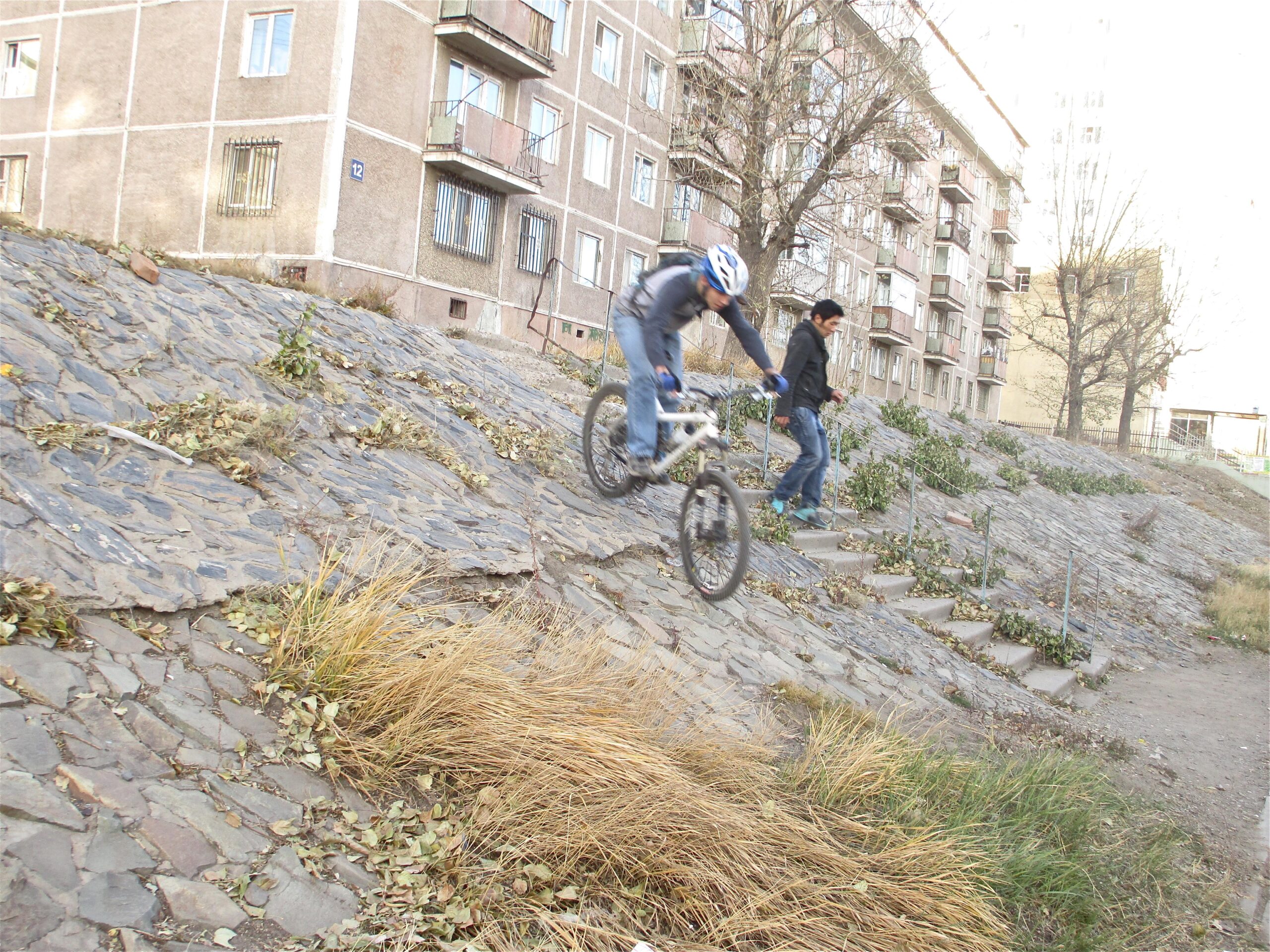 A person on a mountain bike jumps over a rocky slope while another individual walks down nearby stairs, with apartment buildings in the background. The scene is set in an outdoor area with dry grass and fallen leaves. District 15 Network mountain bike trail.