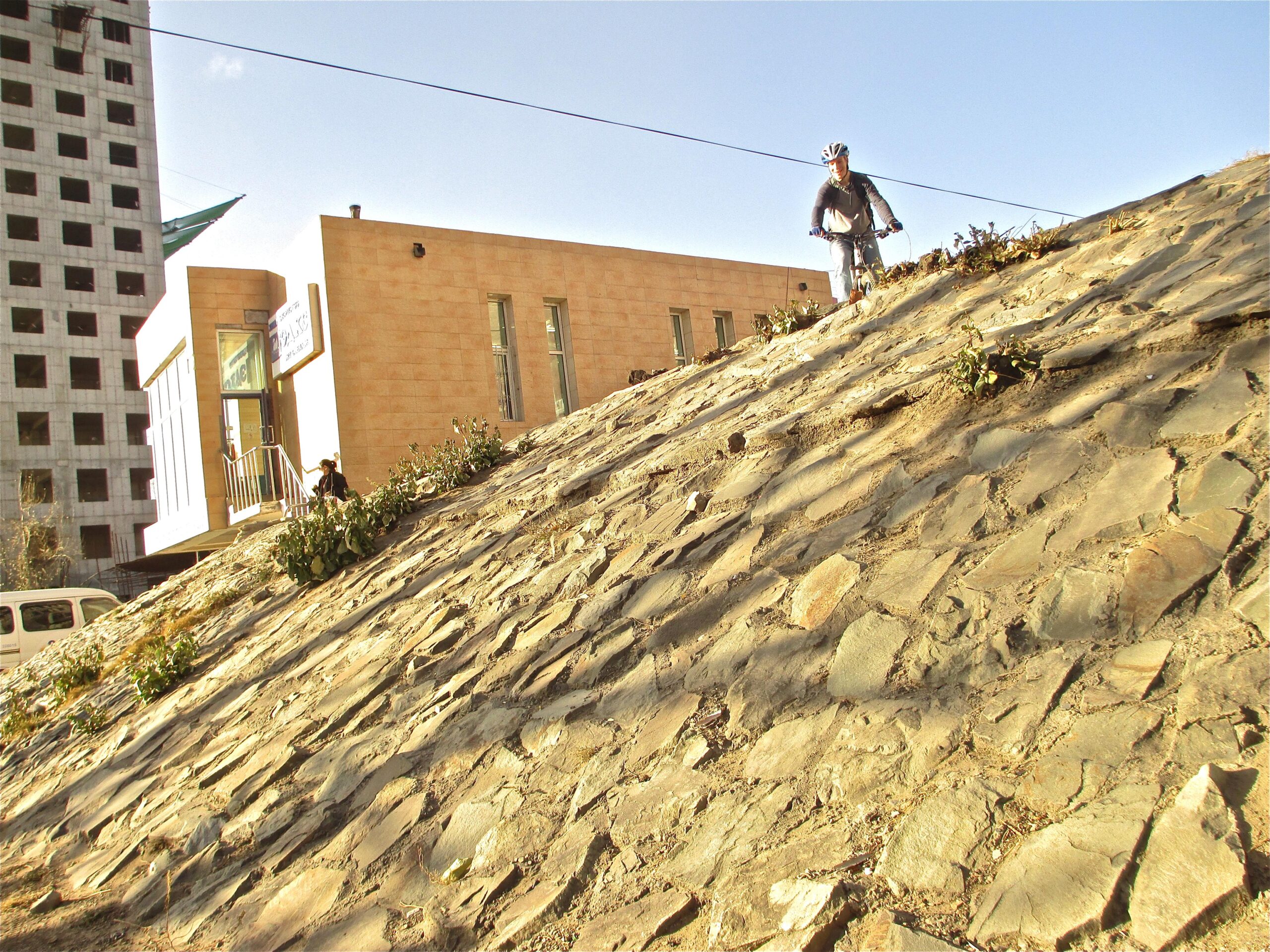 A cyclist riding on a sloped, rocky surface with plants growing in cracks, in front of a modern building. In the background, an unfinished high-rise structure can be seen. The sky is clear and blue, indicating a sunny day. District 15 Network mountain bike trail.