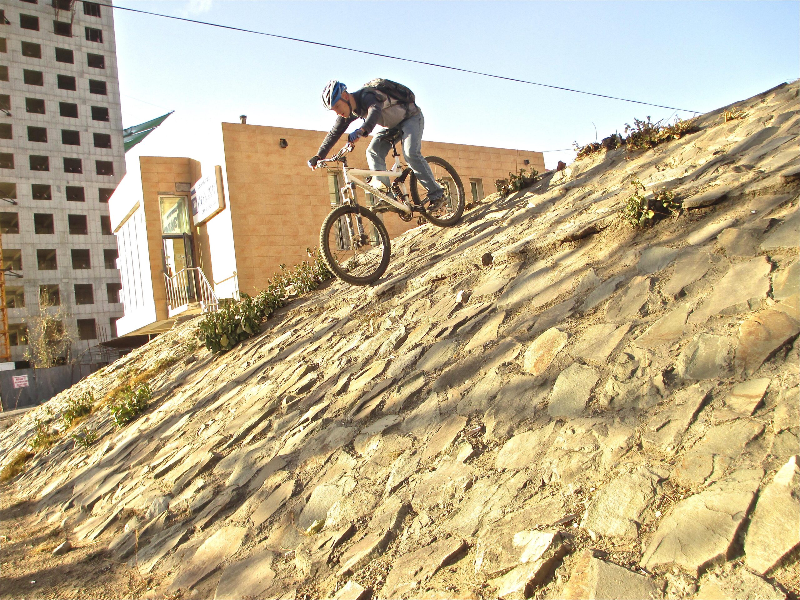 A cyclist jumping down a rocky slope in an urban environment, with a partially constructed building in the background. The cyclist is wearing a helmet and backpack, demonstrating an action-filled moment in mountain biking. District 15 Network mountain bike trail.