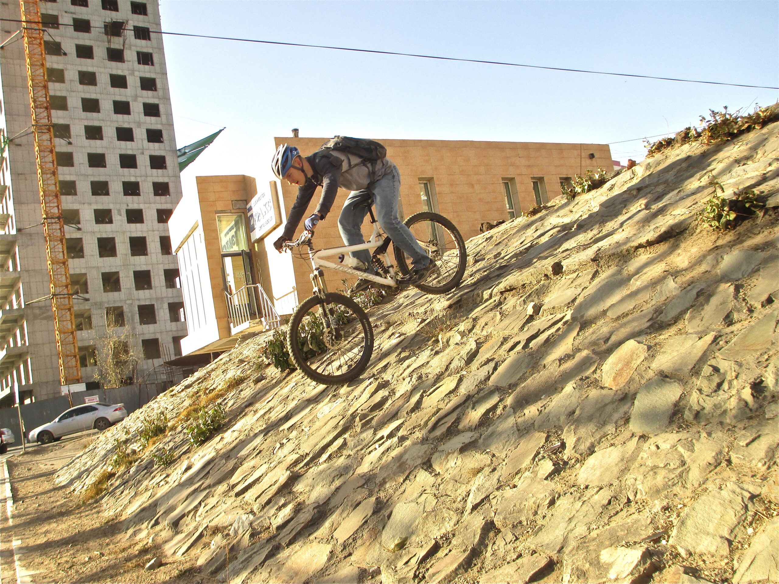 A cyclist in a blue helmet and gray jacket rides a mountain bike down a rocky incline, with a construction site and modern buildings in the background. The scene is set under a clear blue sky, showcasing an urban environment. District 15 Network mountain bike trail.