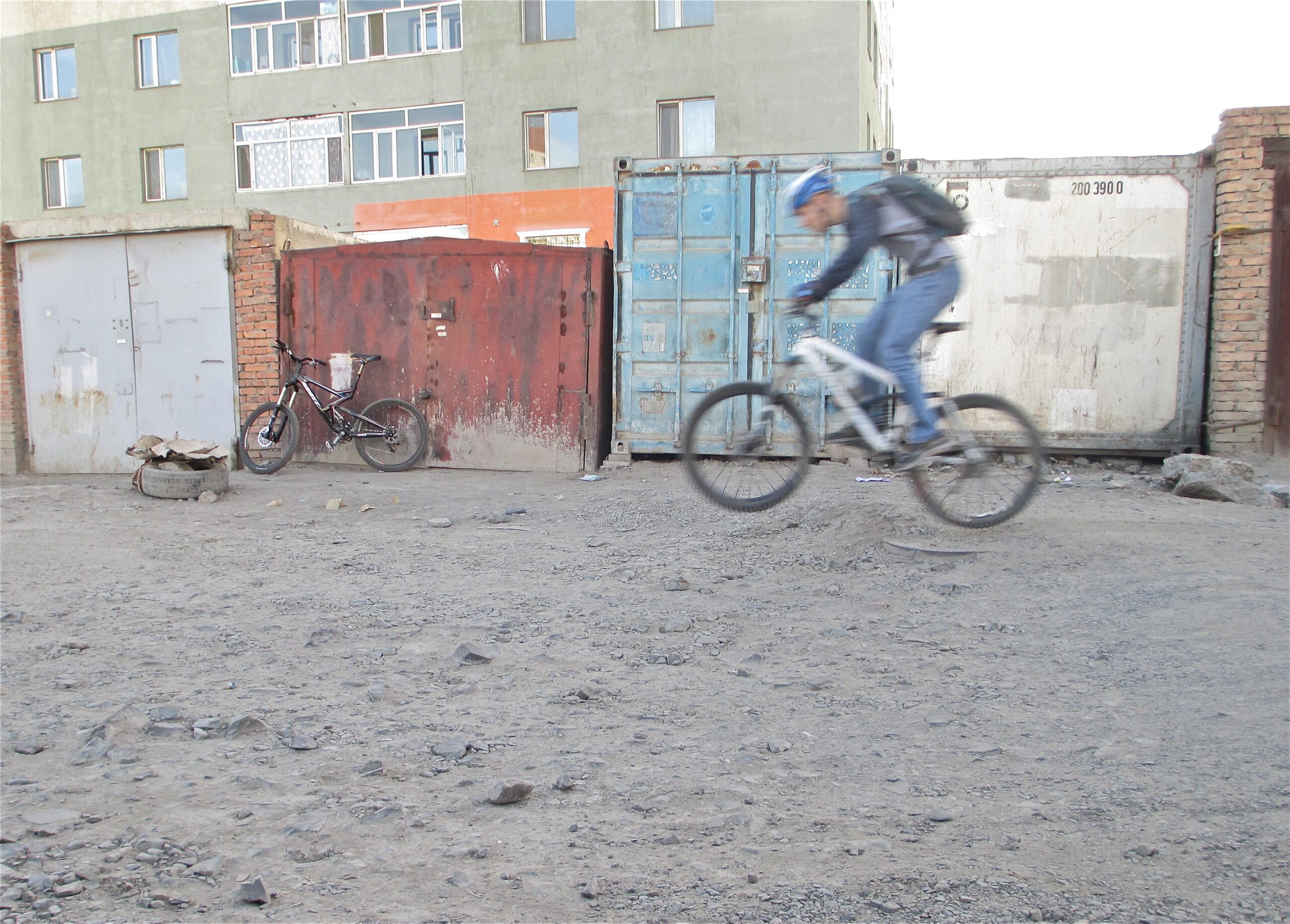 A cyclist in motion rides on a gravel path next to several storage containers and garages. In the foreground, a stationary black bicycle leans against a red metal door, while the background features a building with large windows. The ground is uneven with visible rocks and debris. District 15 Network mountain bike trail.