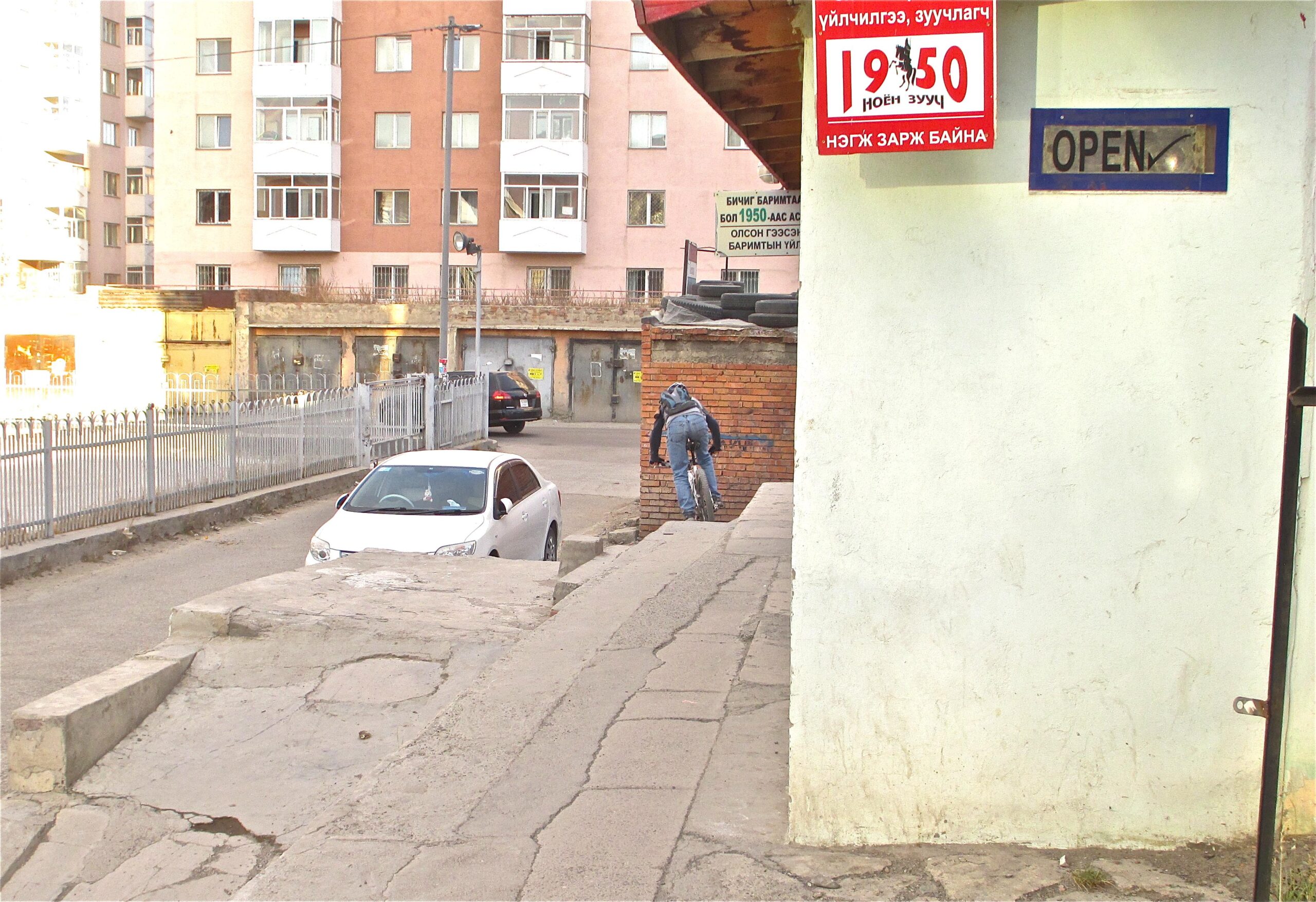 A person riding a bicycle along a narrow walkway beside a building. In the background, apartment buildings and a white car parked on a street are visible. A sign indicating "OPEN" is mounted on a wall to the right, along with another sign displaying a time. The scene captures an urban environment with multiple textures and elements. District 15 Network mountain bike trail.