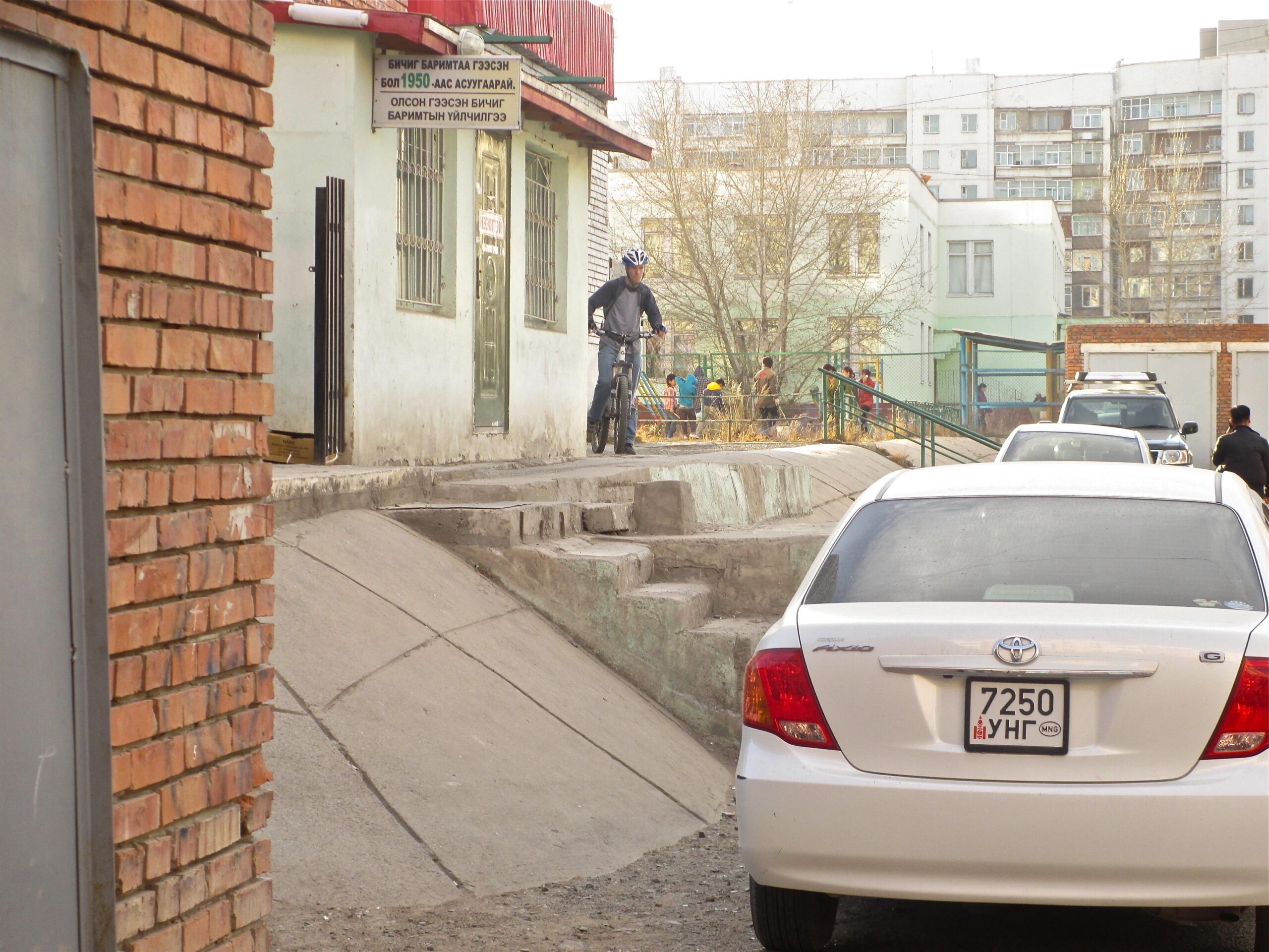 A cyclist riding a bicycle on a ramp beside a building, with a white car parked on the street in the foreground. The background shows residential buildings and people on a playground. A sign in Mongolian is visible on the wall near the entrance of the building. The setting appears to be an urban area. District 15 Network mountain bike trail.