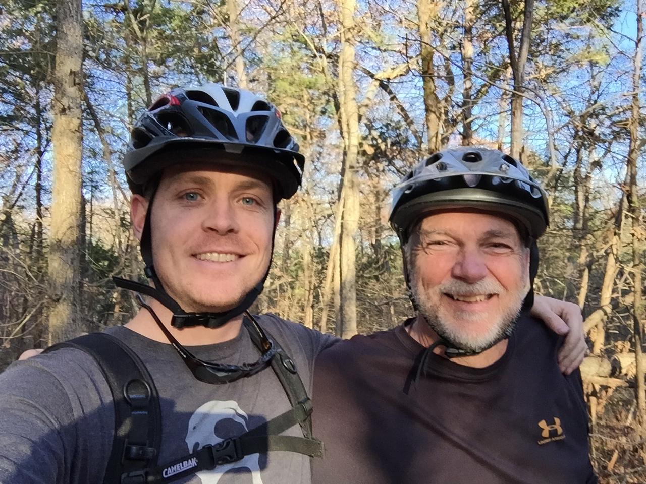 Kona Kahuna: Two men smiling for a selfie while wearing bicycle helmets, standing together in a wooded area with trees in the background. The younger man is wearing a dark t-shirt and a harness, while the older man is dressed in a dark shirt. Both appear to be enjoying a day of outdoor biking.