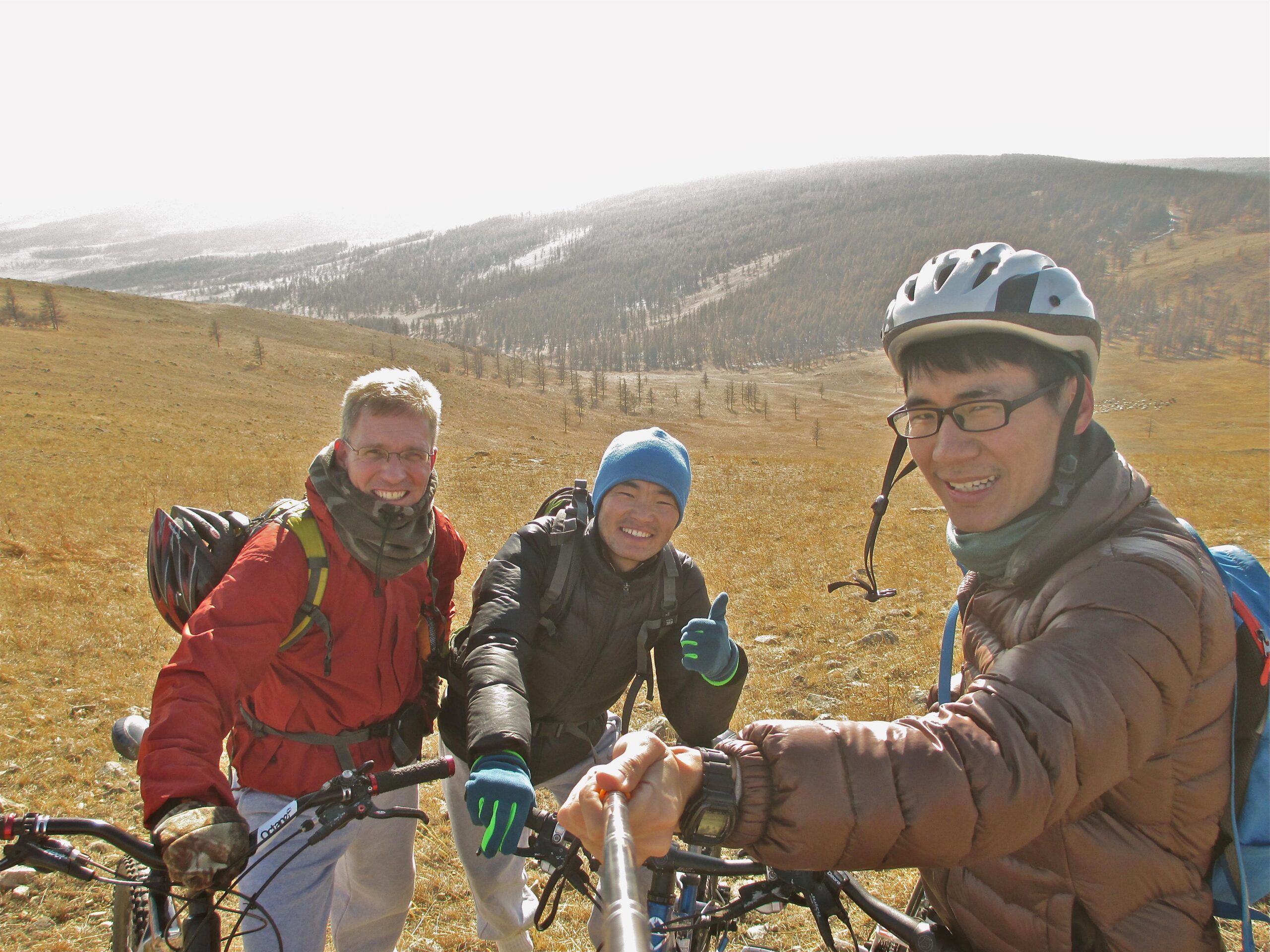 Three cyclists pose for a selfie on a sunny day in a mountainous landscape. They are wearing biking gear and helmets, with two bicycles visible in the foreground. The background features rolling hills and sparse trees, indicating an outdoor adventure. One cyclist gives a thumbs-up, showcasing a cheerful atmosphere. Elst Uul mountain bike trail.